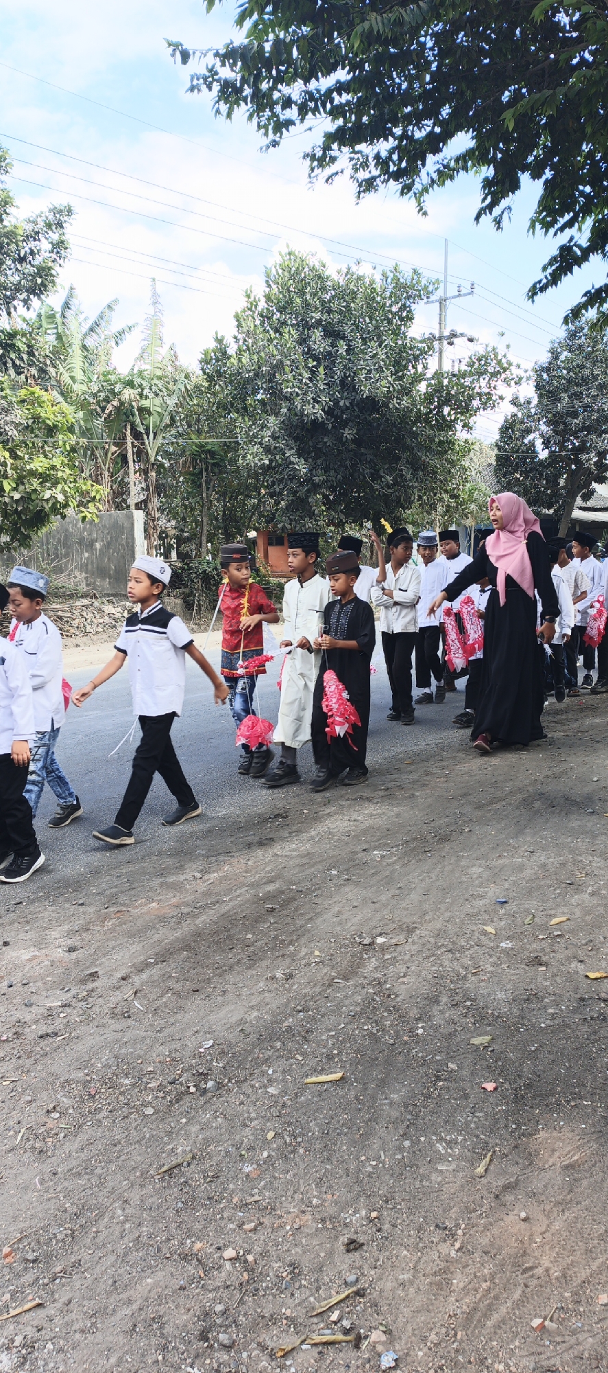 A group of Muslim boys in traditional attire and a woman in a hijab are walking down a street in Grenden, Indonesia. The boys are carrying decorative paper flowers. The street appears to be unpaved and lined with trees and some buildings. The sky is clear and blue.