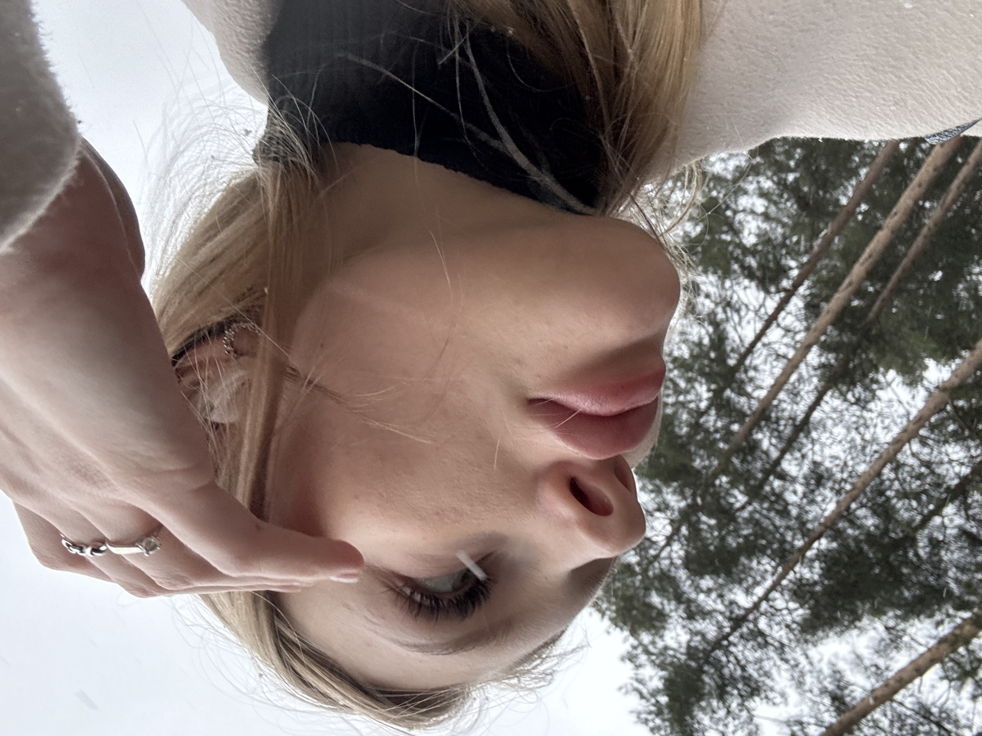 A close-up, low-angle shot depicts a fair-skinned individual, likely female, with blonde hair. The camera is positioned below the subject, looking upwards. The subject's face is prominent, with focus on their eye, nose, and lips. They wear a black garment around their neck and a light-colored coat or sweater. The subject's hand is near their face, adorned with a ring. The background includes trees with bare branches against a cloudy sky. The location is unknown and the city of Perm, Russia can not be verified by solely viewing the image.