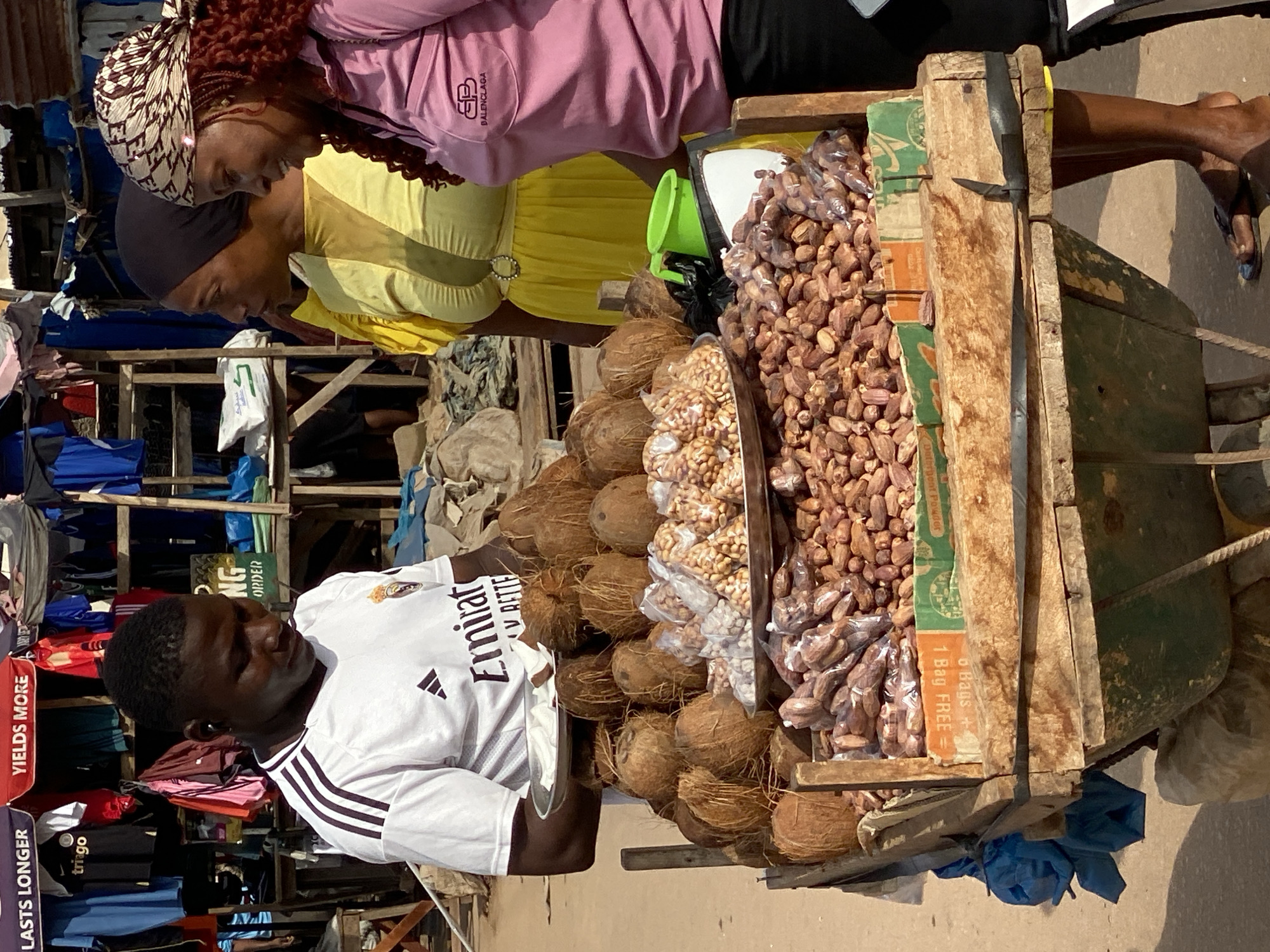 In Sabon Tasha, Nigeria, a vendor stands behind a wooden cart laden with goods. He wears a white Adidas t-shirt with the "Emirates" logo and holds a plate of sliced coconuts. The cart displays a variety of products, including whole coconuts, bagged nuts, and what appear to be dates or dried fruits. Two women are positioned to the vendor's right. One, wearing a yellow dress and headscarf, looks away from the camera. The other, in a pink shirt and a patterned headscarf, smiles. The background reveals a bustling market scene with stalls displaying clothing and other merchandise.