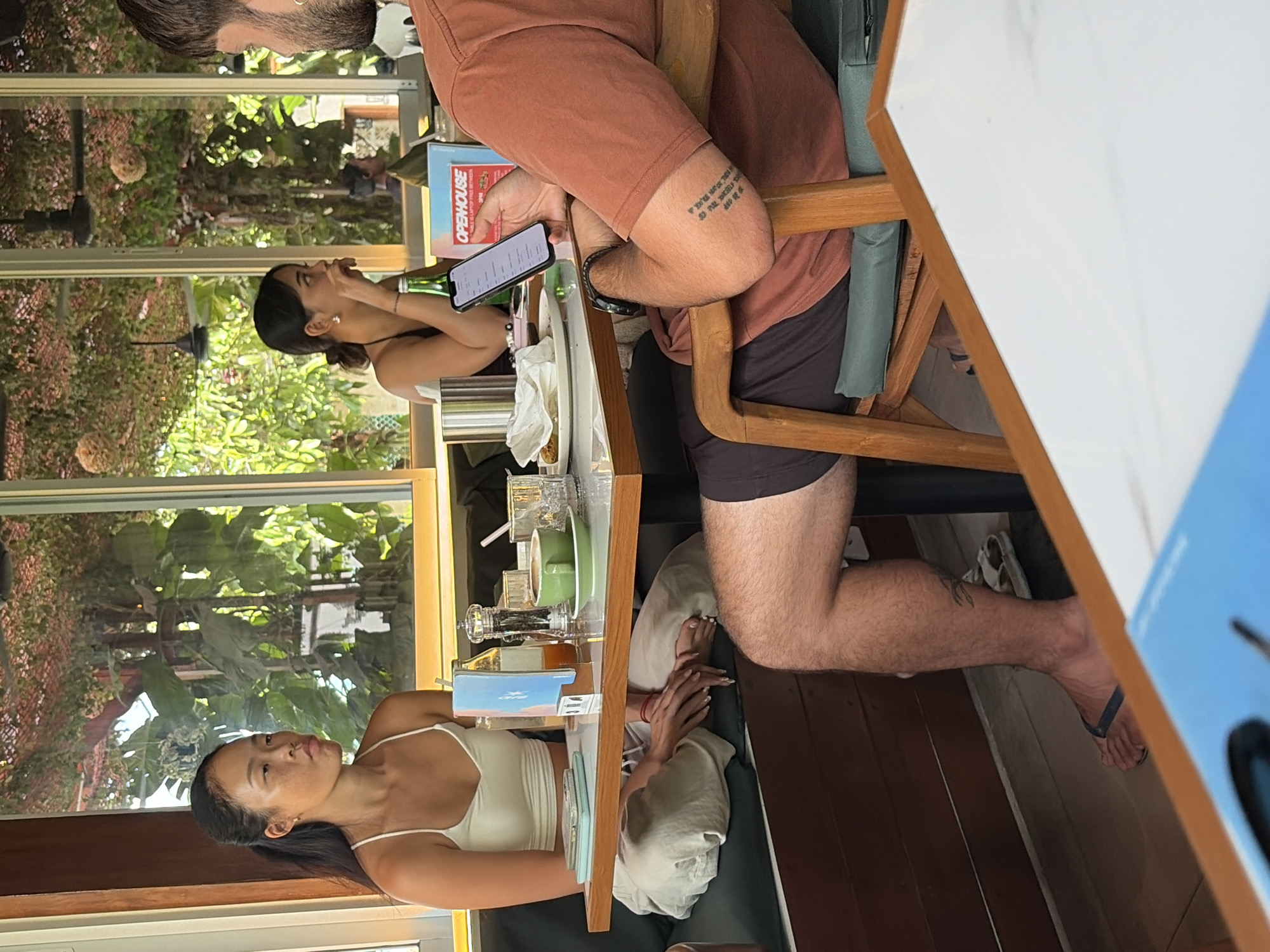 In Mengwi, Indonesia, two women and a man are seated at a table in an outdoor cafe. The woman on the left, with black hair pulled back in a ponytail, wears a white tank top and light-colored pants. She is looking to her left with a neutral expression. Beside her, a green cup and saucer are on the table, along with salt and pepper shakers, a bottle of juice, and a small decorative item.
The second woman, in the background, has dark hair tied back and is wearing a black top. She is also looking down at something on the table, with her hand near her mouth. A green bottle is visible near her.
The man on the right, in the foreground, has dark hair and a beard. He is wearing an orange t-shirt and dark shorts. He is holding a smartphone in his right hand, looking down at the screen. A tattoo is visible on his right forearm. His left leg is extended, and he is wearing a dark sandal.
The cafe appears to be set in a lush environment, with large windows showing greenery and trees outside. The tables are made of light-colored wood, and the seating consists of dark cushions. There are also decorative elements like string lights or hanging plants visible. The overall atmosphere suggests a casual dining experience in a tropical setting.