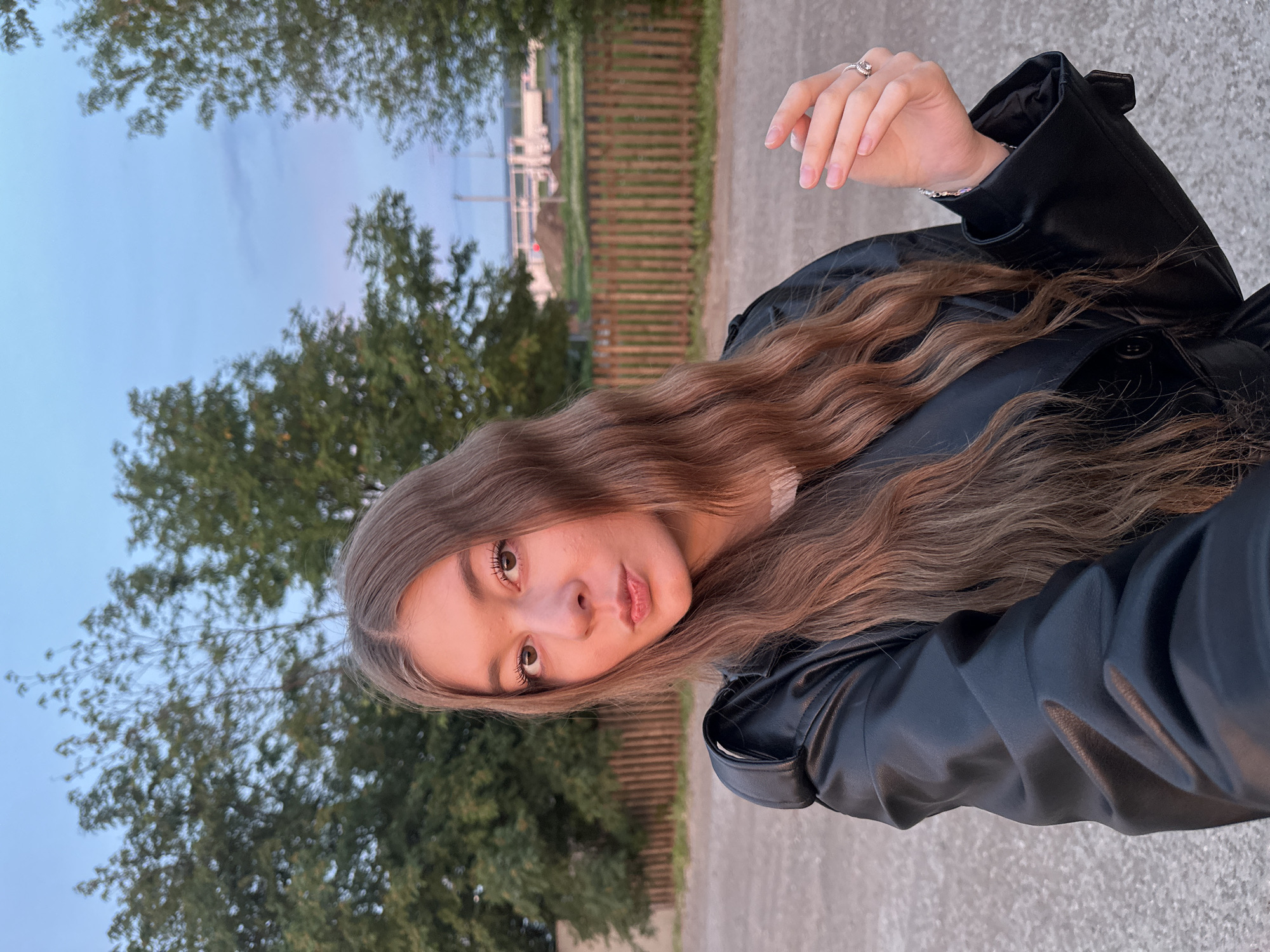 A young woman poses for a selfie in Кузембетьево, Russia. She has long, wavy brown hair and is wearing a black leather jacket. Her hand is raised, displaying a ring. Behind her are trees, a wooden fence, and a glimpse of structures in the distance. The sky is a mix of blue and pale orange, suggesting either dawn or dusk.