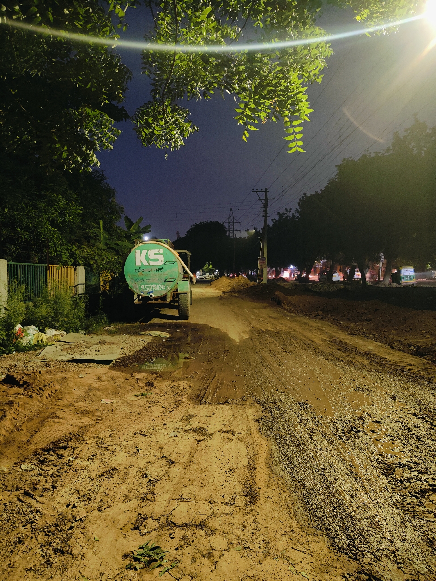 The image captures a scene at night on a partially constructed dirt road in Gurgaon, India. The road is uneven and muddy, with tire tracks visible. On the left side of the road, a green water tanker truck is parked. The tanker has "KS" written on it in white letters, along with some text in Hindi and a phone number. Next to the truck, there is a green metal fence and overgrown green vegetation. Piles of debris and garbage are also present near the fence. In the background, trees line the road, and streetlights illuminate the area, casting a warm glow. The sky is dark, indicating it is nighttime. Power lines and poles are visible against the sky, and some distant lights suggest a populated area or ongoing construction work. The overall impression is one of an active construction site at night, with basic infrastructure visible.