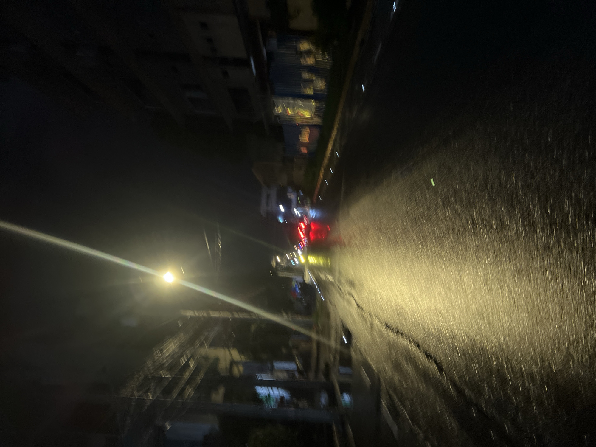 The image depicts a nighttime view of a street in Davao City, Philippines. The perspective is slightly tilted, with buildings visible on either side of the street. Rain or wet conditions are indicated by the streaks of light reflected on the road surface. Streetlights illuminate the scene, casting a bright glow. In the distance, there are car headlights and tail lights, suggesting vehicular traffic. A construction site or scaffolding is apparent on the left side of the road. The environment is dark, and details are somewhat obscured by motion blur and the weather conditions.