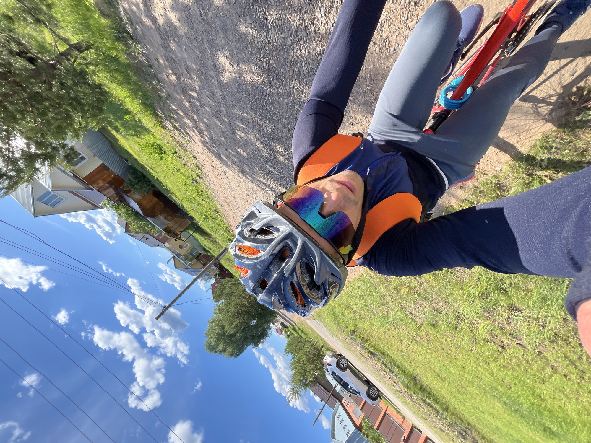 A cyclist is on a bike on a gravel path in Chaikovsky, Russia. The cyclist wears a helmet, cycling glasses, a blue and orange jersey, dark blue sleeves, and light gray pants. A red bicycle frame is visible, along with a blue coiled lock. The cyclist is facing the camera, possibly taking a selfie. To the left of the cyclist, several houses and telephone poles with lines are visible under a blue sky with white clouds. To the right of the cyclist, there is a grassy area, a white car, and buildings.