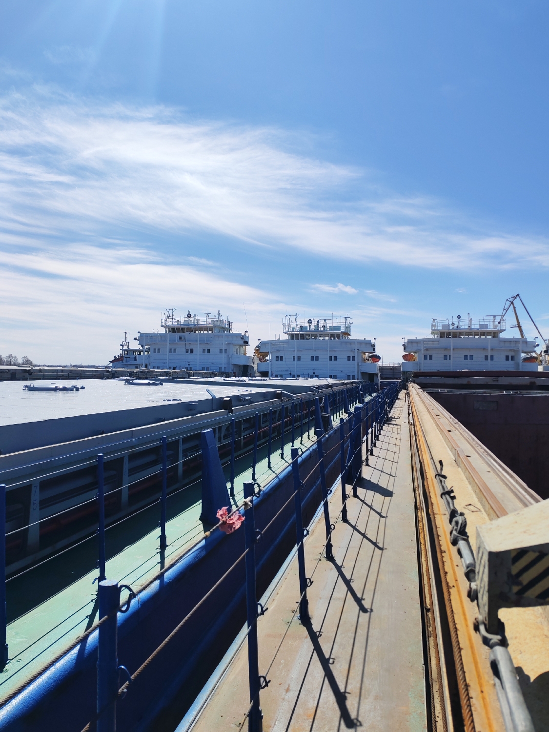 The image depicts an industrial maritime scene at what appears to be a port or shipyard, consistent with the location context of Rybinsk, Russia. The sky is bright blue with scattered, wispy white clouds, suggesting clear weather conditions during the daytime, likely morning or early afternoon given the distinct, elongated shadows.
In the background, three large, white river vessels, possibly cargo barges or ships, are prominently docked. They are aligned horizontally and feature multi-deck superstructures with numerous windows and various pieces of equipment, including antennae, on their roofs. A crane arm is visible extending from the stern of the vessel on the far right.
In the foreground, a narrow walkway or gangway stretches from the lower right towards the middle-left of the frame. It is bordered on the left by a robust blue metal safety railing, which casts long, sharp shadows across the walkway. Below this railing, a blue trough-like structure and a green-painted hull or surface are visible. A small piece of red and white fabric or rope is tied to the blue railing near the center. On the right side of the walkway, a heavy, rusty yellow metal beam or rail runs parallel to the path, equipped with cables and a concrete block marked with black and yellow warning stripes.
No people are visible in the image, and the scene conveys a sense of calm, suggesting the vessels are either moored, undergoing maintenance, or awaiting activity. Faint numbers "7 5" are discernible on the green hull surface below the blue railing on the left.
