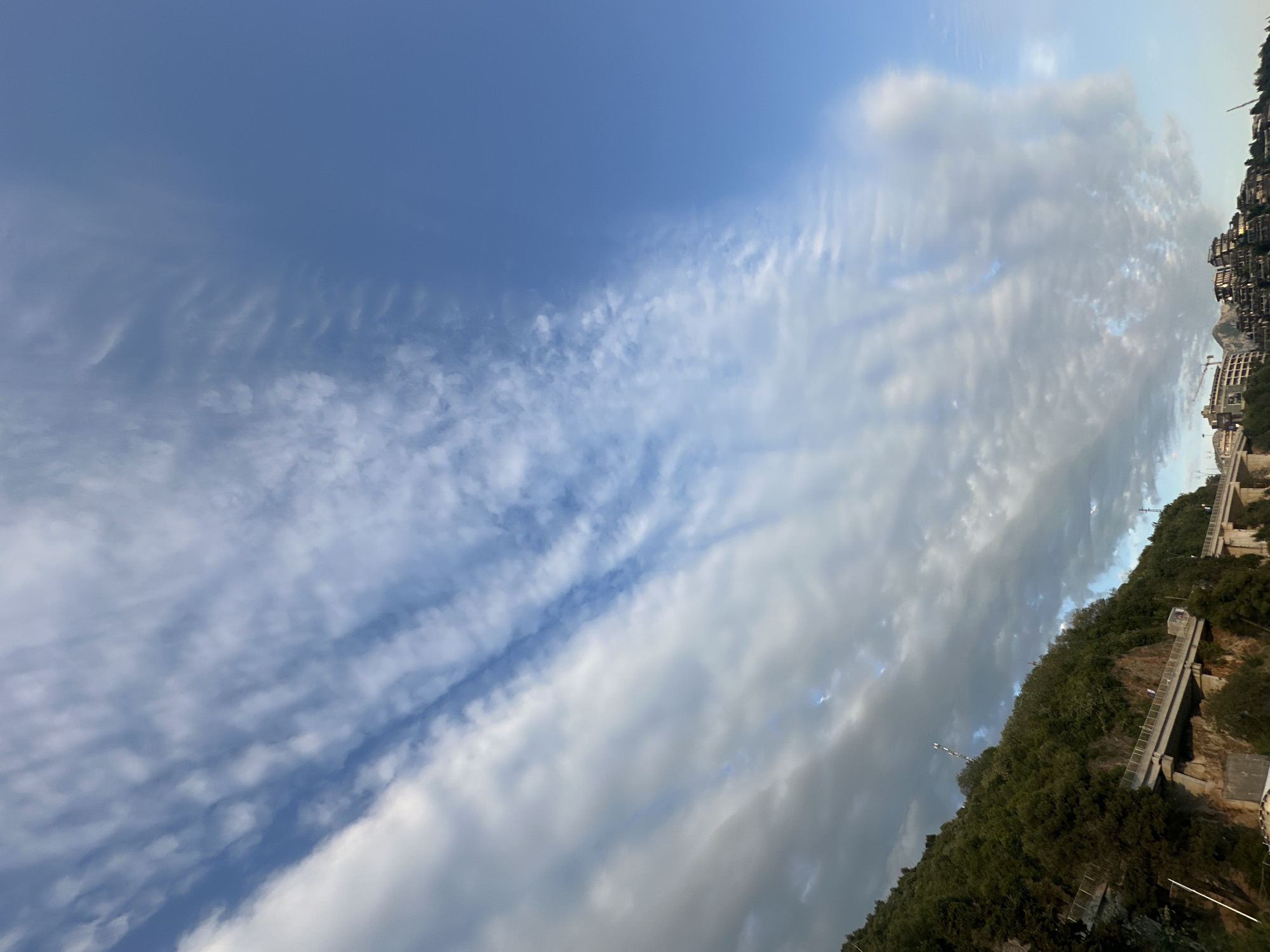 The image, when correctly oriented, presents an expansive daytime landscape dominated by a dynamic sky above a hilly coastal area. The upper and left portions of the frame are filled with a vibrant blue sky interspersed with diverse cloud formations. On the left, thin, wispy clouds stretch across the blue, forming delicate, parallel streaks, reminiscent of cirrus or altocumulus undulatus. Towards the right, the clouds become denser and lumpier, displaying shades of grey and white, indicative of thicker altocumulus or stratocumulus clouds, with some clear blue patches peeking through.
Below the sky, the landscape features a steep, verdant hillside covered in dense green trees and vegetation. Clustered along the slope are numerous modern multi-story buildings, likely residential or hotel structures, built into the terrain. Infrastructure such as concrete pathways or long sets of stairs ascend parts of the hill, and a construction crane is visible near some of the buildings, suggesting ongoing development.
The bright, natural lighting indicates it is daytime. The varied cloud cover suggests a partially cloudy but bright day. The combination of steep, vegetated hillsides and densely packed modern architecture points to a coastal urban environment, consistent with Budva, Montenegro. There are no people visible, and no discernible text in the image.