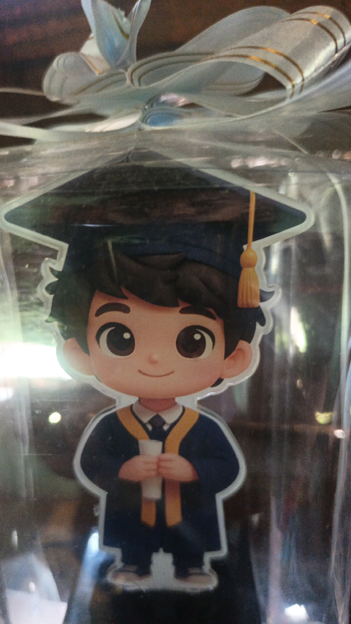 The image shows a close-up of a graduation-themed decoration, possibly an acrylic standee or a gift topper. The main subject is a cartoonish figure of a young boy wearing a graduation cap and gown. He has dark hair, large, expressive eyes, and a slight smile. He is holding a scroll or diploma in his hands. The graduation cap is dark blue with a yellow tassel. The gown is also dark blue with yellow trim. The figure has a white outline. The background is blurry and appears to be indoors, with a brown, possibly wooden surface visible at the top and a darker, reflective surface below. There is also a light blue ribbon with gold accents at the very top of the image, suggesting it is part of a package. The overall lighting is dim, with some light reflections visible on the protective casing of the decoration. There is no visible indication of the city of Sungai Rasau, Indonesia, or any specific location.