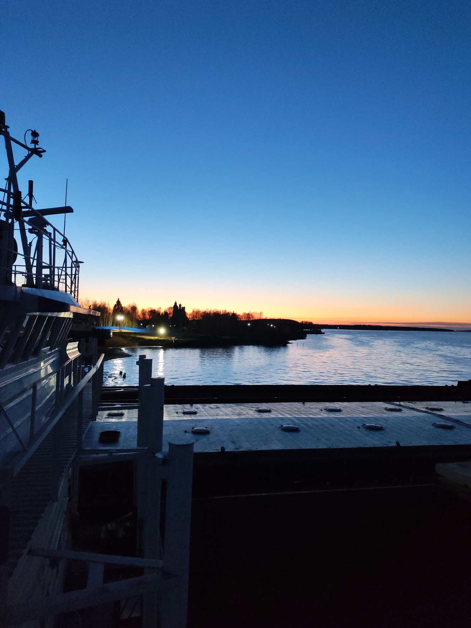 The image captures a serene twilight scene from the perspective of a ship or large vessel. The sky dominates the upper portion, displaying a gradient from deep blue at the zenith to warmer orange and yellow hues at the horizon, indicative of either sunrise or sunset. The water below is calm, reflecting the soft light of the sky and the artificial lights from the distant shore.
In the immediate foreground on the left, the dark silhouette of the vessel's superstructure, including railings, a mast, and what appears to be a radar dome and other navigational equipment, frames the view. To the right, part of the ship's deck is visible, featuring several oval-shaped hatches or access points.
The middle ground is dominated by a wide expanse of water. On the distant shoreline, a line of bare trees is silhouetted against the glowing horizon. Several bright artificial lights illuminate a section of the land, casting distinct reflections onto the water. Further along the shore, the dark, recognizable silhouette of a building with multiple spires, possibly a church or a castle, stands out against the colorful sky.
The overall atmosphere is one of peaceful quietness. No people are visible, and there is no discernible text in the image. The scene suggests the vessel is either docked or anchored in a large body of water, such as a river, lake, or bay.