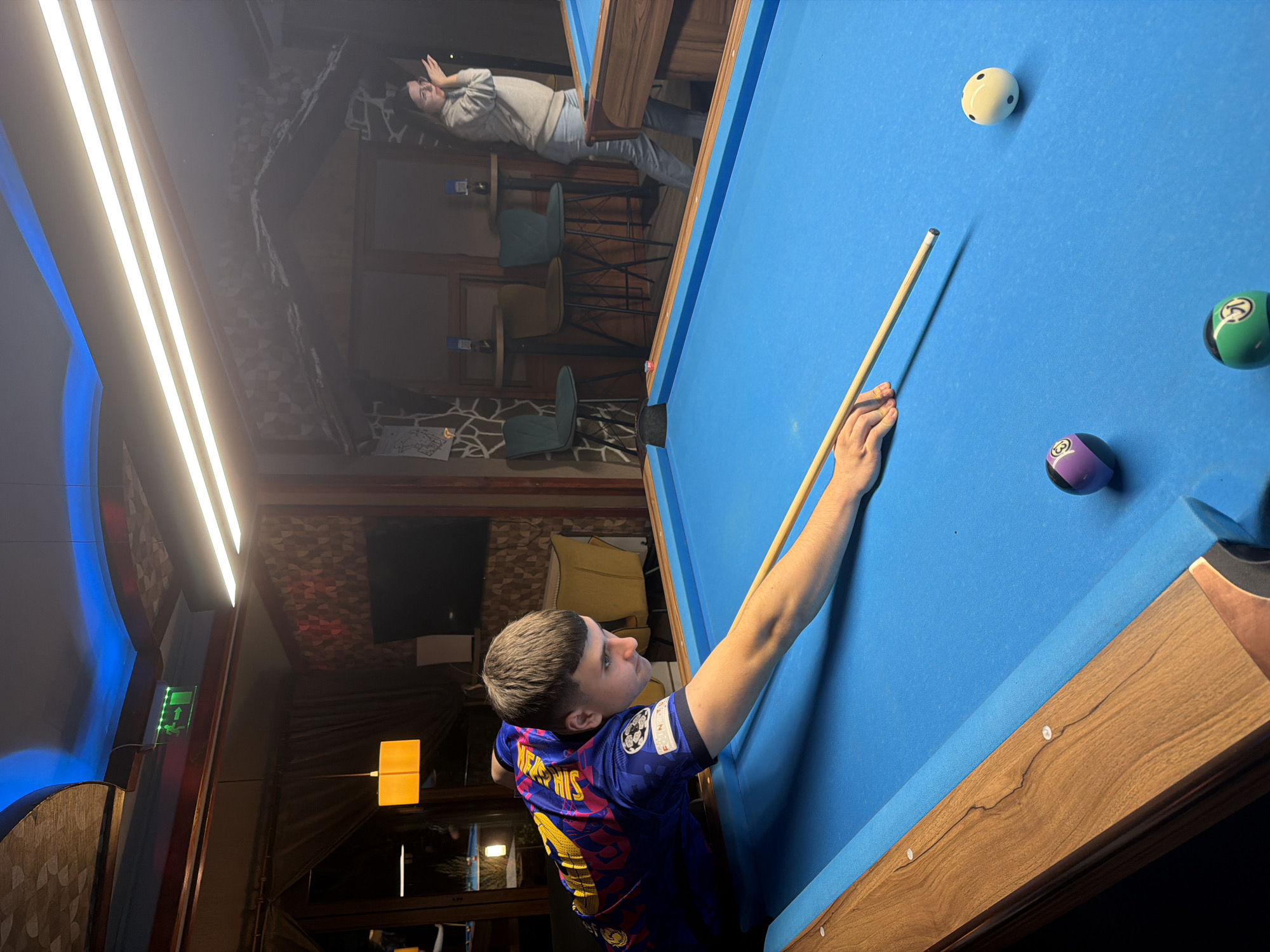 A young man in a Barcelona soccer jersey is playing pool in a room with a wooden-themed interior. He's aiming the cue stick towards a white cue ball, with a purple 9-ball and a green 3-ball nearby on the blue pool table. In the background, a person wearing a light-colored sweater stands near a high table and chairs. The location is Plovdiv, Bulgaria.