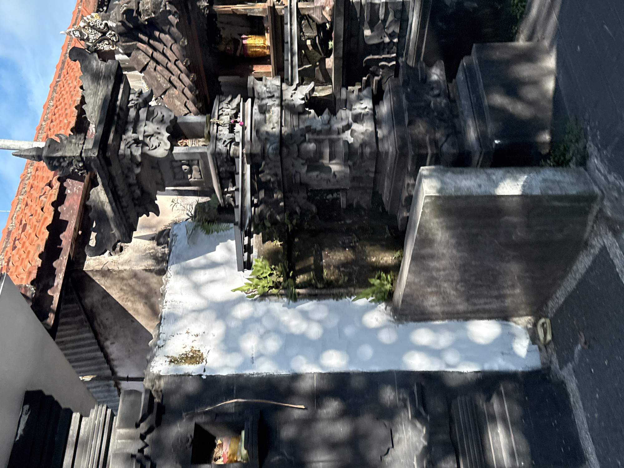 This image captures a close-up view of a traditional Balinese temple or building structure, rich in architectural detail, under bright daylight in Kuta Selatan, Indonesia. The scene is dominated by dark gray and black stone, meticulously carved with intricate traditional motifs, forming various tiered platforms and ornate gate-like elements.
On the left side, a vibrant terracotta tiled roof slopes downward, adorned with decorative wooden or stone finials and ridge ornaments, one of which features elaborate swirling designs and a white accent. A slender, pointed spire rises from one of the structures. Within a darker recess of the stone architecture, a subtle gleam suggests a golden or bronze object, possibly an offering or shrine detail.
Below the main stone structures, a white-painted wall or ledge is illuminated by dappled sunlight, creating striking patterns of light and shadow, indicating the presence of overhead foliage. Small green plants and foliage emerge from crevices and grow on the ledges, adding touches of nature to the stone. The ground appears to be a dark, paved surface, also exhibiting shadow patterns. A clear blue sky is visible in the background, suggesting fair weather. There are no people visible, and no specific activities are taking place. No text is visible in the image.