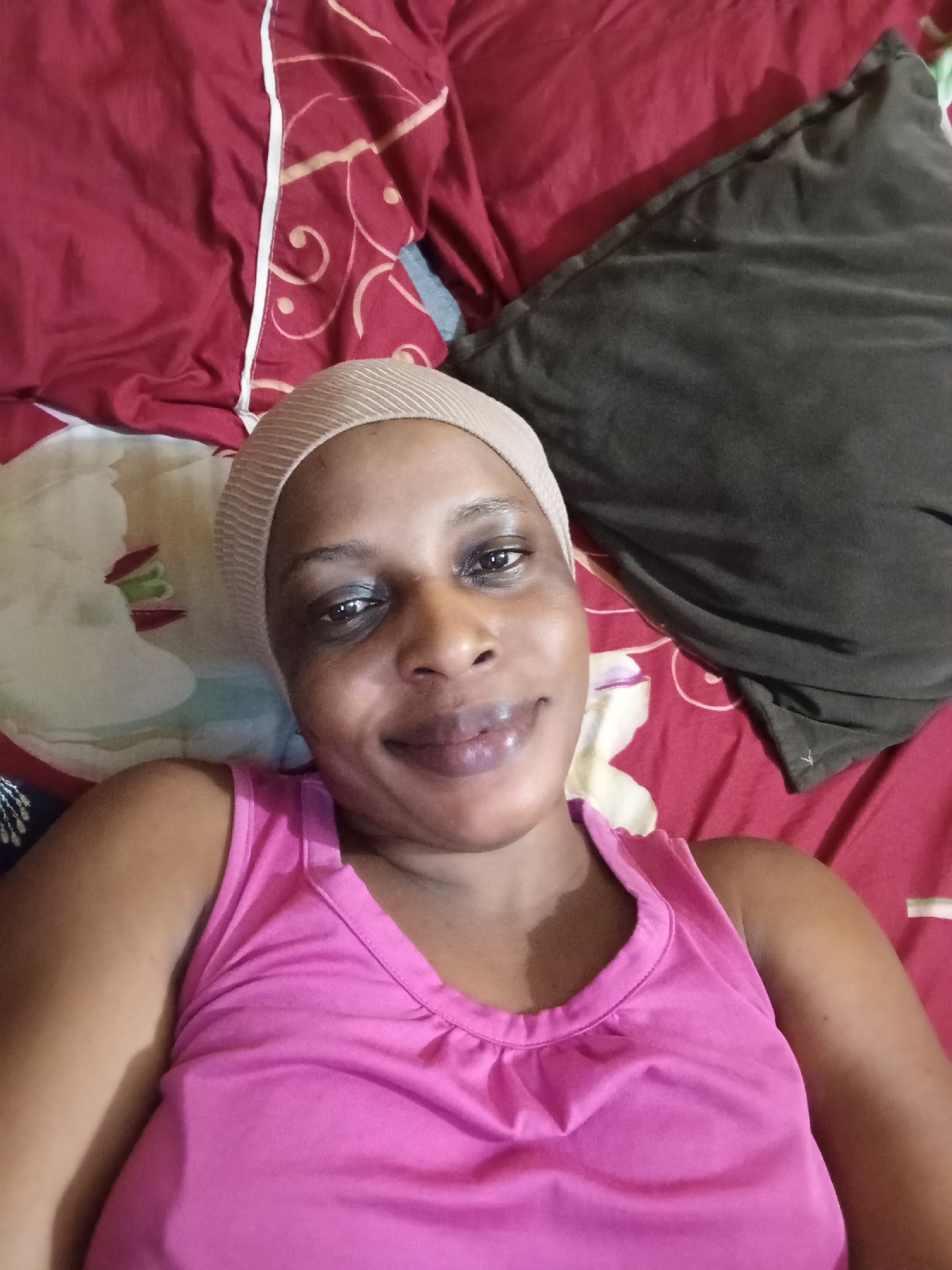 A woman is lying on a bed in what appears to be an indoor setting. She is wearing a pink sleeveless top and a beige head covering. The bed is covered with patterned red and white sheets and has a dark gray pillow. It's not possible to confirm if the location is Narayi, Nigeria without additional information.