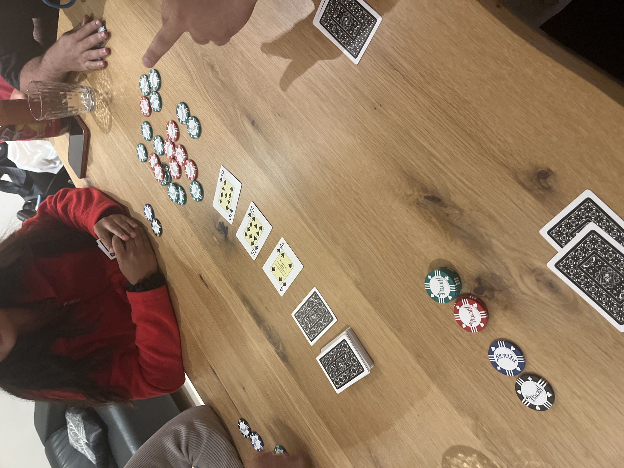 The image shows a group of people playing a card game on a wooden table. Several poker chips of various colors (green, red, blue, white) are scattered across the table. Playing cards are arranged in different configurations, including face up and face down piles. Some cards display suit and number values, such as 9 and 10 of spades. Hands are visible as are people in the periphery of the image. There is no indication of location.