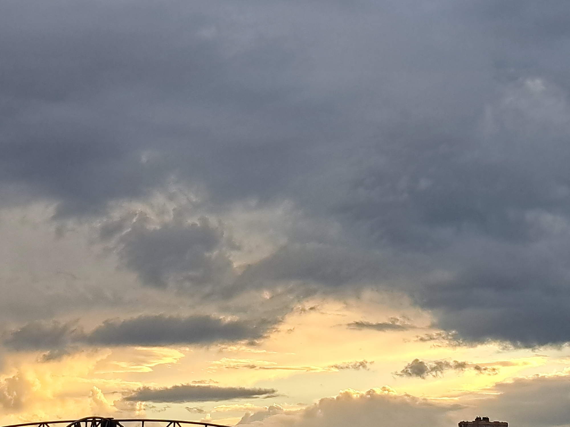 This is a photograph of a cloudy sky over the city of Irkutsk, Russia. The sky is filled with a mix of dark gray storm clouds and lighter, golden clouds illuminated by the setting sun. In the foreground, the silhouette of a bridge railing is visible against the dramatic sky. The overall impression is one of an approaching storm or the aftermath of one, with a striking interplay of light and shadow in the clouds.