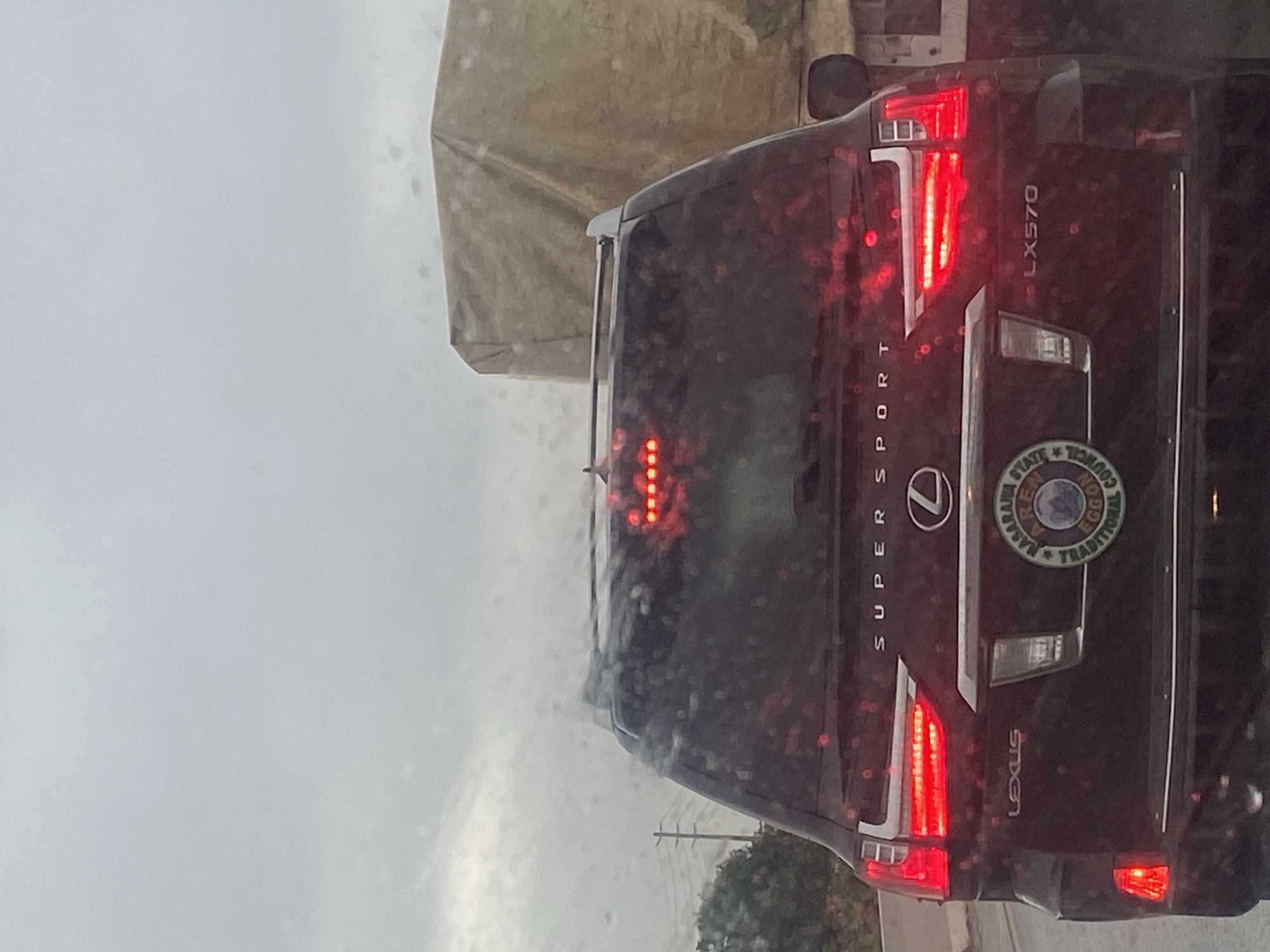 The image shows the rear of a black Lexus LX570, a luxury SUV, on a road. The vehicle has its brake lights illuminated, indicating it is likely stopped or slowing down. The "SUPER SPORT" badge is visible on the tailgate, along with the Lexus logo. Below the logo, there is a circular emblem that appears to be a seal or crest, with text that includes "NASARAWA STATE", "EGGON TRADITIONAL COUNCIL". The license plate is not clearly visible. To the right of the Lexus, a portion of a large truck with a tan tarp covering its cargo is visible. The sky is overcast, and there are visible water droplets on the car's rear windshield, suggesting rain. The overall scene suggests a cloudy day in a traffic or roadside environment. The presence of "NASARAWA STATE" on the emblem indicates the vehicle is likely in Nasarawa State, Nigeria, and the reference to "EGGON TRADITIONAL COUNCIL" further narrows the potential location within the state. Keffi is a prominent city in Nasarawa State, and it is plausible this scene is within or near Keffi.