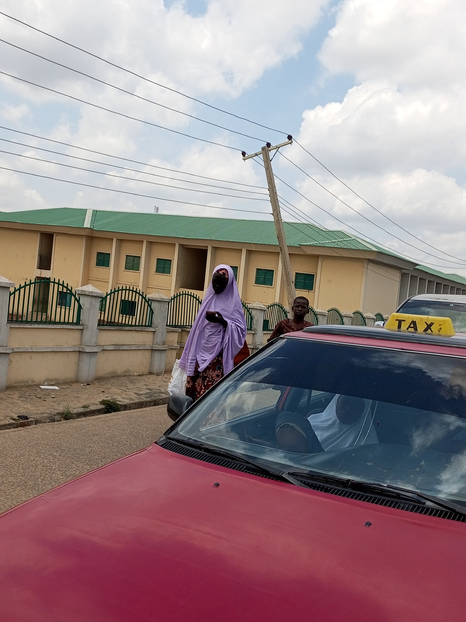 In Pantame, Nigeria, a red car is parked on a street with a paved surface. A woman wearing a purple hijab and a patterned dress stands beside the car, holding a plastic bag. To her right, a young boy in a brown shirt is also visible. A yellow taxi sign is mounted on the roof of the red car. In the background, a light yellow building with a green roof and green-framed windows is situated behind a green metal fence. Power lines and a utility pole are present in the sky.