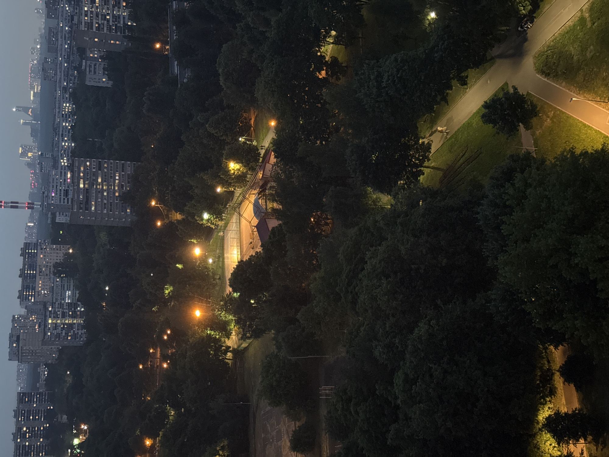 The image depicts a nighttime view of a city park adjacent to a residential area. In the foreground, dense, dark green trees fill much of the frame, with some interspersed patches of illuminated grass. A paved pathway winds through the park, with a lamppost illuminating a section of it. Two people are visible walking on the pathway in the mid-ground. Further back, a playground or sports complex is lit by artificial lights, with a fence surrounding it. Beyond the park, a cluster of tall apartment buildings are visible, their windows lit, suggesting an urban environment. In the far distance, the city skyline is visible, with more buildings and what appears to be a tall, red and white striped structure. The overall impression is of a moderately populated urban area at night, with a clear separation between a green, park-like space and the built environment of the city.