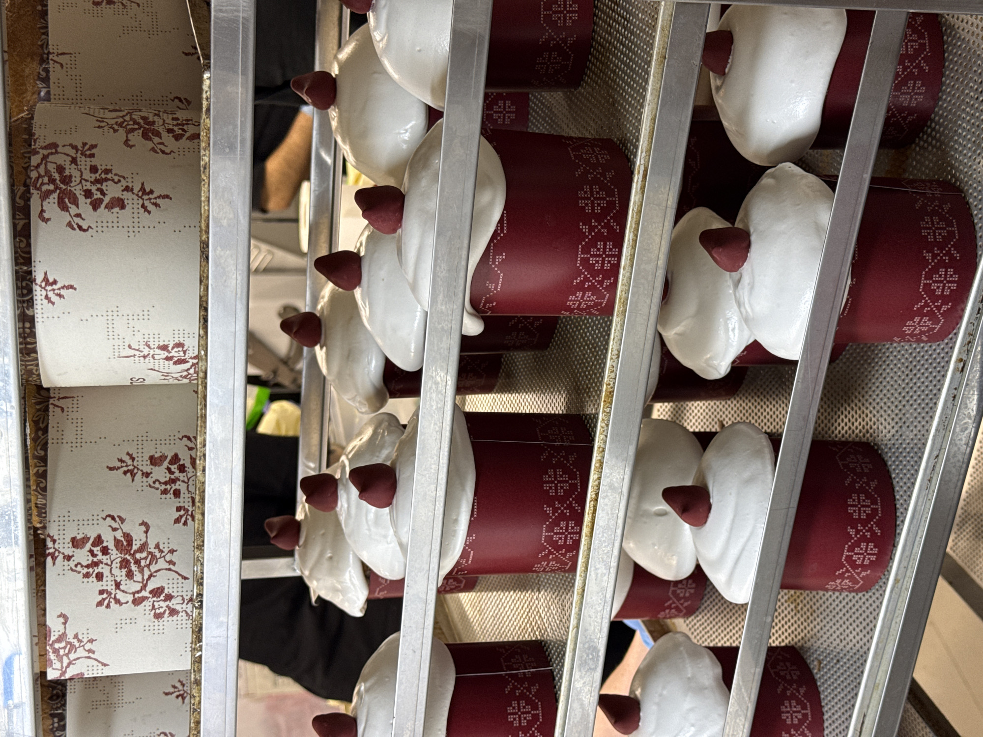 The image shows several cakes in a metal rack. The cakes are red with a white frosted top and a red decorative piece on top. The rack also holds tubes of decorative paper. The scene is likely in a bakery or food preparation area. The exact location within the city of Belgrade, Serbia is not discernible from the image.