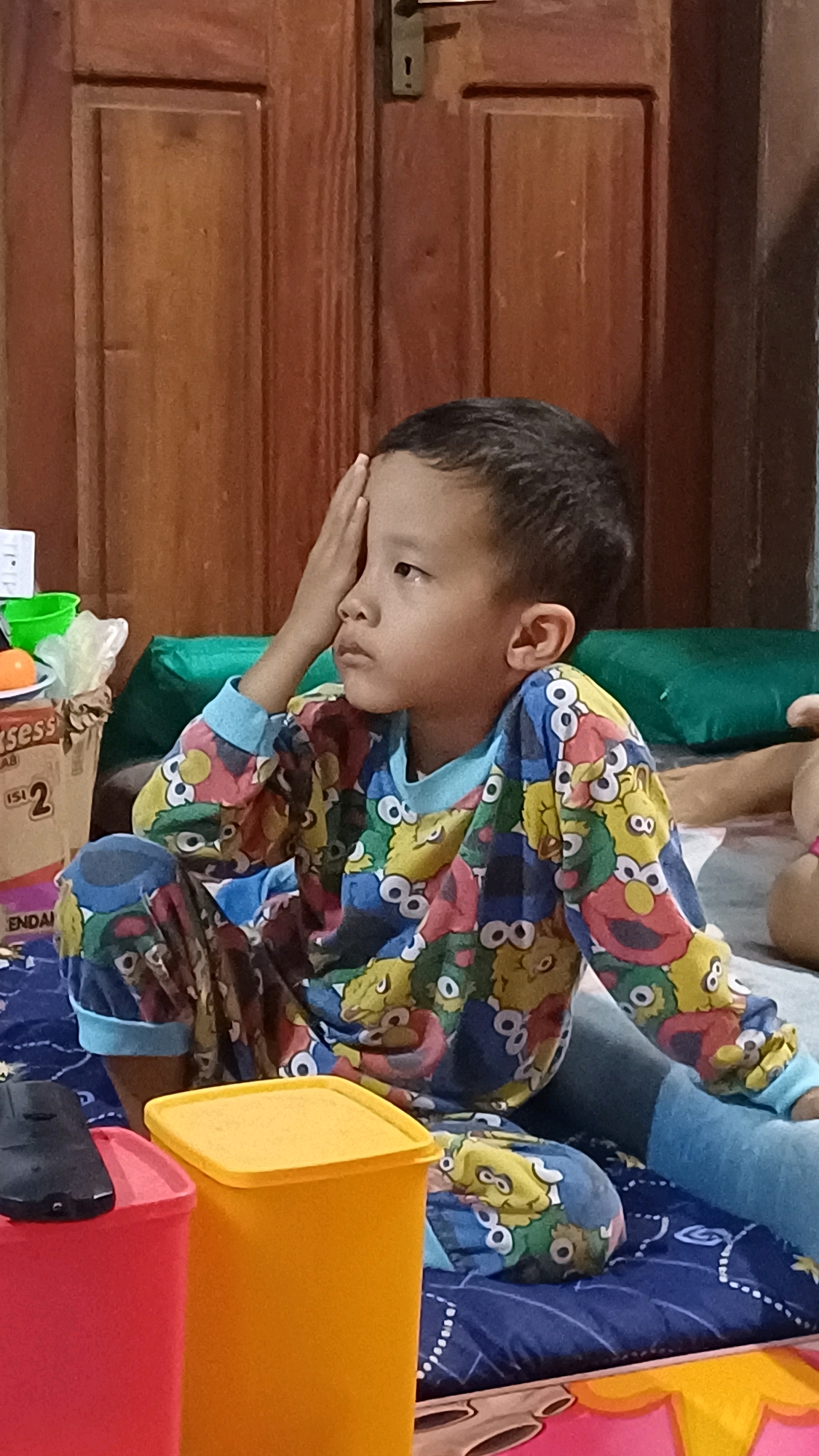 The image depicts a young boy, likely of Southeast Asian descent, sitting on a blue patterned mat. He is wearing pajamas adorned with cartoon characters, predominantly yellow and blue with red and green accents. His right hand is raised, resting on his forehead, as he gazes to his left with a pensive expression. In the foreground, to the left, are two plastic containers, one red and the other yellow. Behind the boy, a wooden door with a dark metal handle and lock is visible, suggesting an indoor setting. The presence of a green pillow behind him indicates he is in a relaxed or casual environment. The scene appears to be an interior residential space. The image does not contain enough information to determine a specific location within Wonokerso, Indonesia, beyond it being an indoor scene.