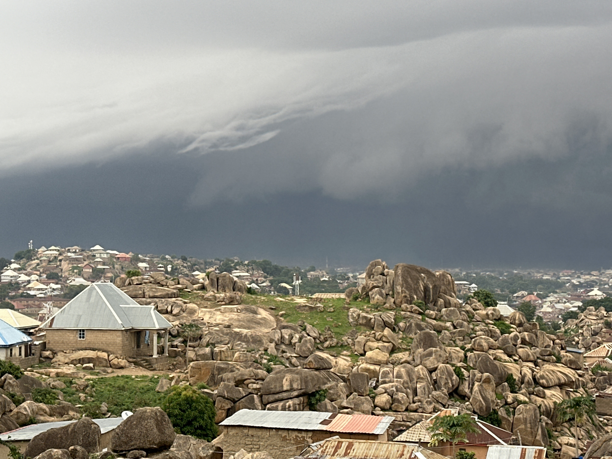 The image shows a landscape in Jos, Nigeria, characterized by rocky terrain and buildings interspersed among the rocks. The foreground features various buildings with metal roofs, surrounded by numerous large rocks and sparse vegetation. In the middle ground, there are more buildings spread across the rocky landscape, with some elevated areas. A large grouping of rocks and a white antenna-like structure on a grassy clearing stand out. The background shows a dense urban area with many buildings, fading into a hazy distance. Above the scene, a dark, ominous storm cloud dominates the sky.