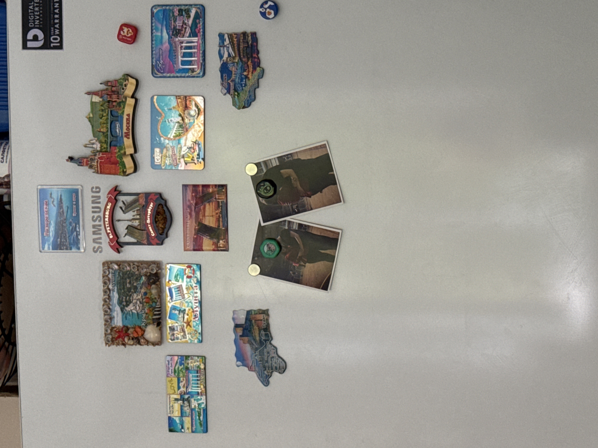 A close-up, eye-level, indoor shot shows a refrigerator door covered with various magnets and two photographs. The background is a plain white refrigerator door. The magnets are colorful and depict landmarks and scenes from various locations, possibly tourist souvenirs. One magnet shows the Moscow Kremlin. Another appears to be a map of Crimea. There are also magnets with text that is partially visible, but appears to be in Russian. The photographs are of a couple embracing, with a small circular green magnet affixed to the top of one. A small red square magnet with a symbol is also visible. There is no indication of the city of Saratov, Russia in this image.