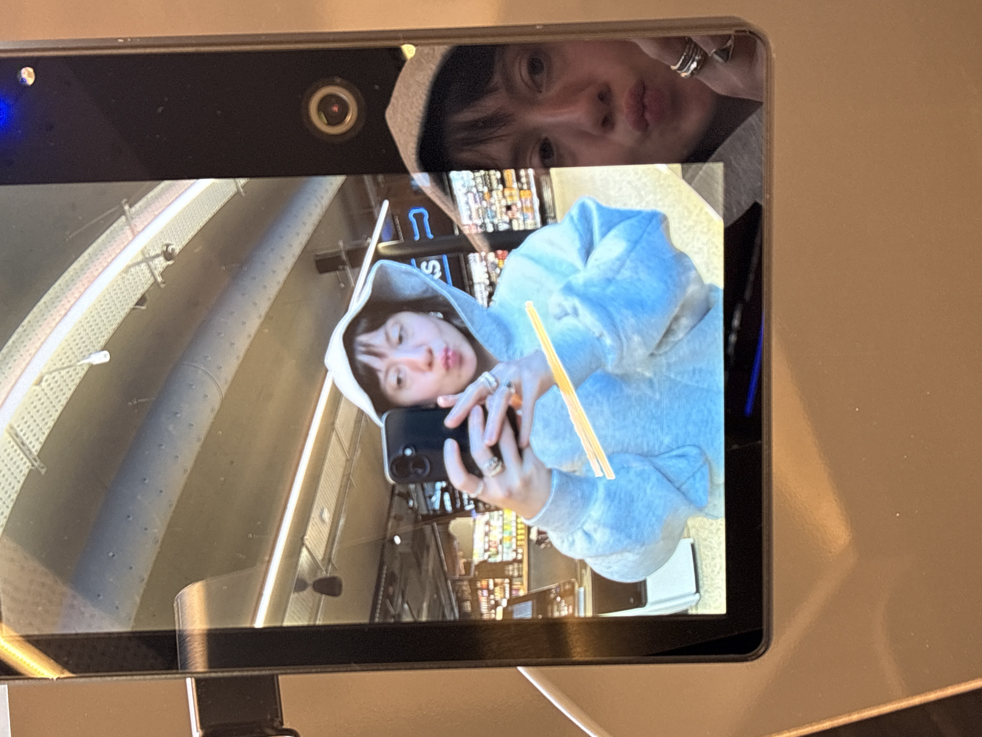 The image shows a screen displaying a selfie of a person wearing a light gray hoodie and several rings, with two yellow objects held in their hand. The background visible in the selfie suggests a supermarket interior with shelves stocked with goods. Above the selfie is a mirrored, inverted reflection of the person's face with an eye-level view, indicating the photo was taken with a device held above. The photo is being viewed on a screen. The camera indicates the presence of other people. The location is unknown.