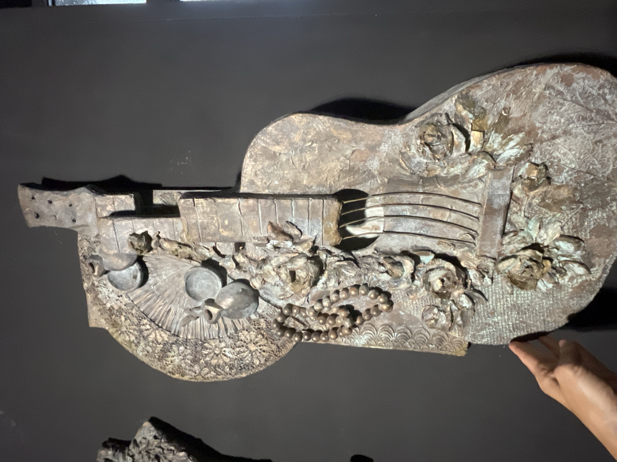 The image shows a guitar relief sculpture mounted on a dark-colored wall. The relief depicts a guitar decorated with floral motifs (roses and leaves), small vases, and a string of beads. The sculpture has a distressed, aged appearance, possibly made of bronze or a similar metal. A hand is partially visible in the lower right corner, suggesting someone may be interacting with the sculpture. Another similar sculpture is located at the bottom of the frame. The picture was taken in Sochi, Russia.