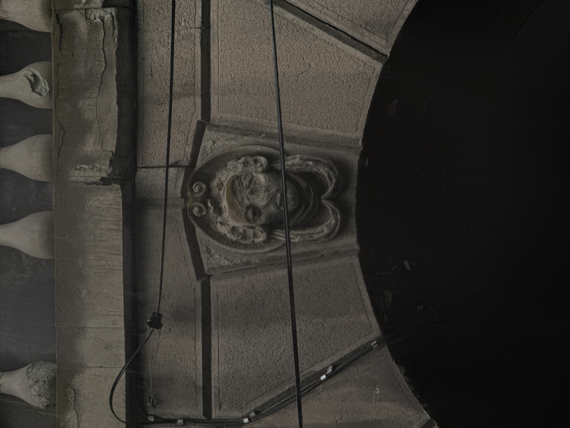 A close-up, exterior shot captures a carved stone face set into an arched architectural feature, likely part of a bridge or building in Tbilisi, Georgia. The face, with its weathered features and ornate hair or headdress, appears in the center of the frame. Above it, a stone balustrade runs horizontally across the top of the image, with several balusters visible, some showing signs of damage. Dark electrical wires are strung across the scene, intersecting the architectural elements and partially obscuring the carved face. The lower portion of the image is dominated by a dark, shadowed archway, suggesting an opening or passage beneath the structure. The overall tone is muted, with a focus on the textures of stone and the stark lines of the wires.