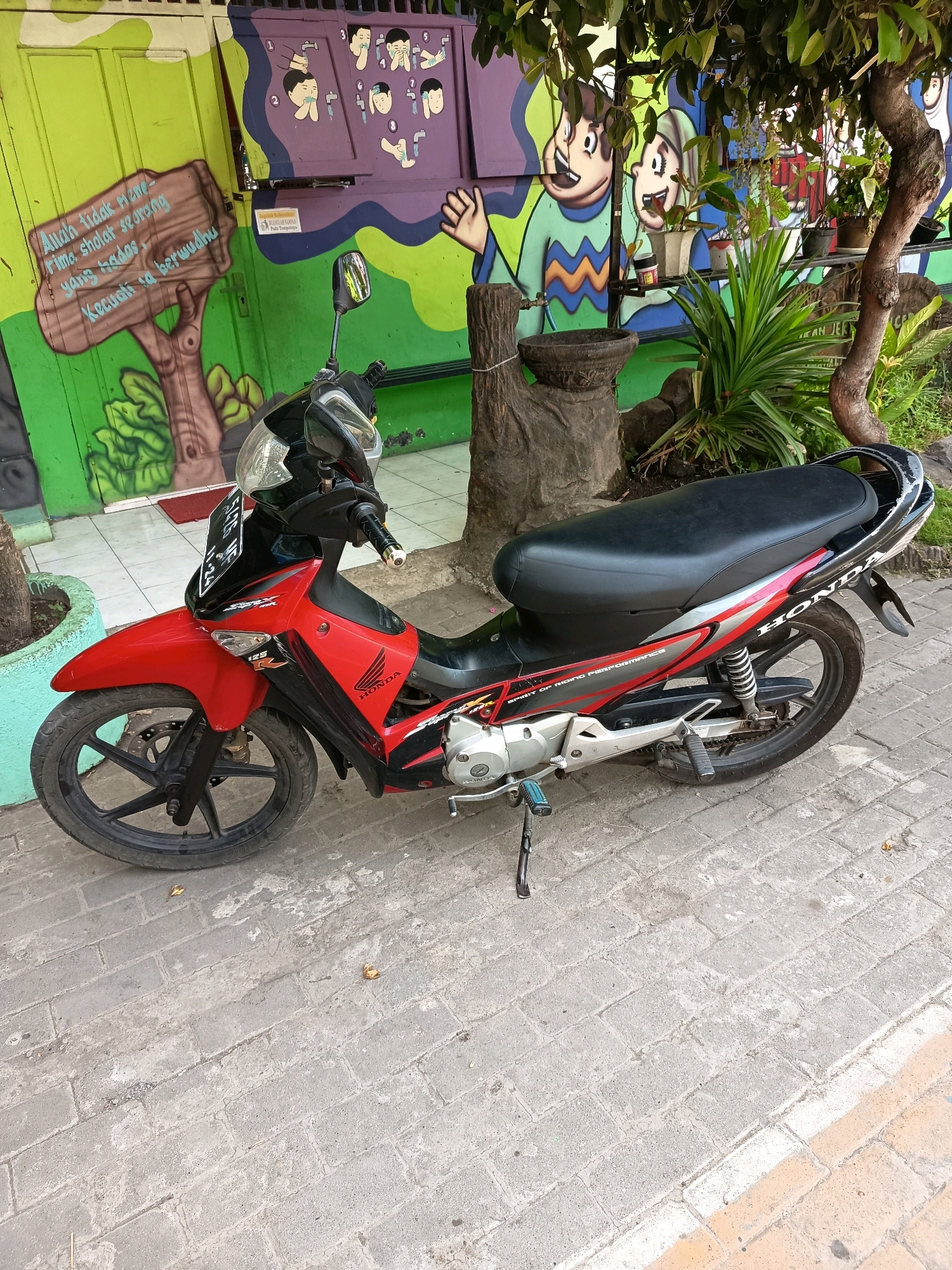A red and black Honda motorcycle is parked on a brick sidewalk in front of a building with colorful murals. The murals depict cartoon characters and Arabic script. To the left of the motorcycle is a green-painted door with a mural of a tree and more Arabic script. To the right of the motorcycle, a tree with green leaves shades the scene. The building appears to be a public facility, possibly a school or community center, given the educational murals. The motorcycle has a license plate that reads "E1428 UE 11-24". The area is outdoors, and the lighting suggests daytime.