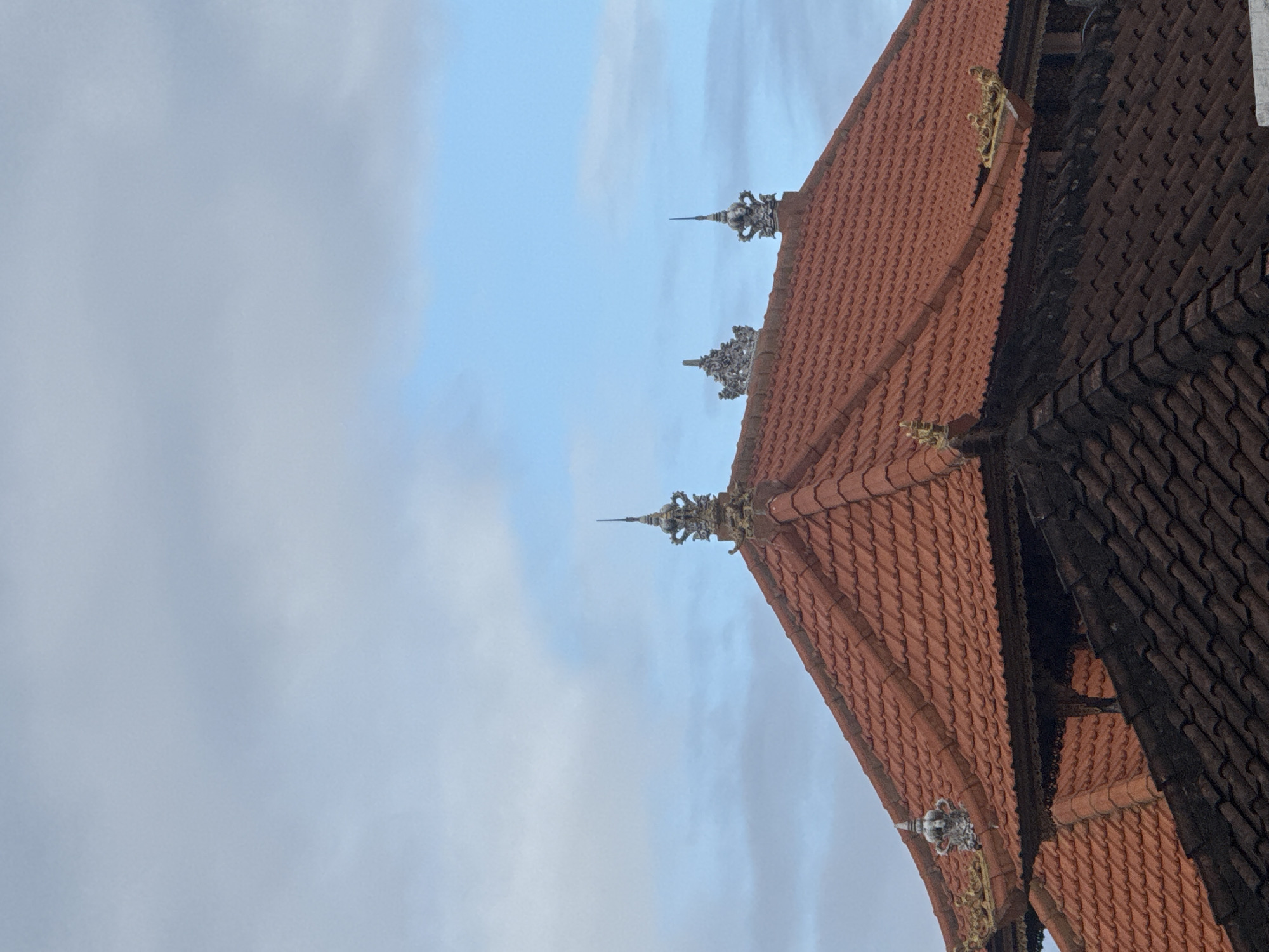 The image shows a close-up of a tiled roof with intricate architectural details against a cloudy sky. The roof tiles are predominantly a reddish-orange color, arranged in a consistent pattern. Along the edge of the roof, there are several decorative elements, appearing to be made of stone or metal. These embellishments are ornate, featuring spiky or crown-like designs, some with a lighter, weathered appearance, and others with gold accents.
The sky is overcast with light, diffused clouds, suggesting either early morning, late afternoon, or a generally cloudy day. The lighting is soft and even, without strong shadows, which is consistent with a cloudy atmosphere. The perspective is from below, looking upwards towards the roof and the sky. The scene is devoid of any people or animals.
The image is dominated by the textures of the roof tiles and the detailed ornamentation. The two different colored sections of the roof tiles (reddish-orange and a darker, almost black color) suggest different materials or age. The dark section on the right appears to have more aged, possibly weathered or soot-covered tiles, contrasting with the brighter red tiles on the left.
The architectural style of the decorative elements hints at a location with a rich cultural heritage, possibly a temple or a traditional building. Given the location context of Kuta, Indonesia, this style could be indicative of Balinese architecture. The overall mood is serene and perhaps slightly mysterious due to the ornate but aged details and the subdued lighting. There is no visible text in the image.