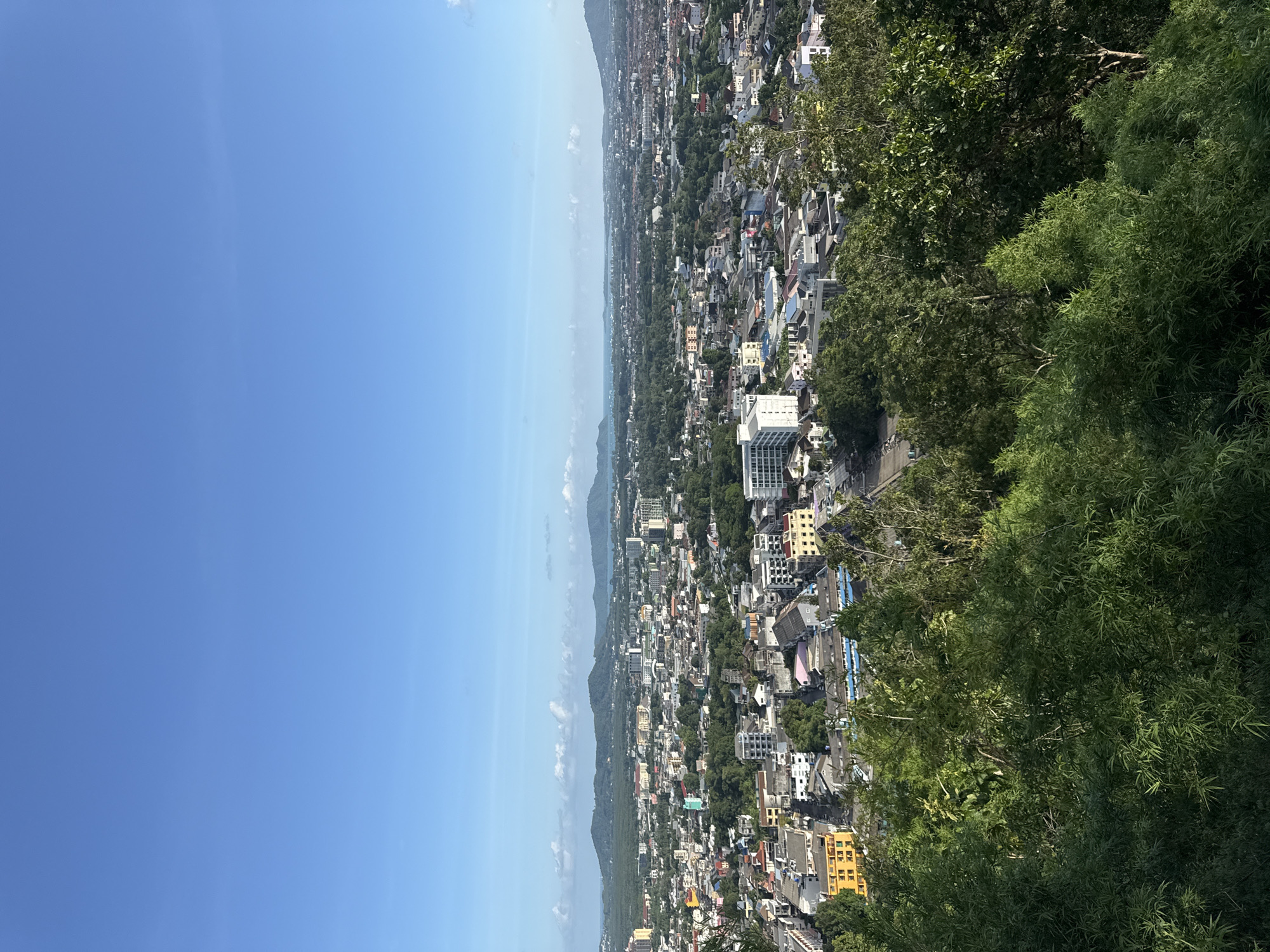 An aerial, wide-angle shot captures a sprawling cityscape nestled between lush green hills and the ocean under a clear blue sky. The city, identified as Ratsada, Thailand, is densely packed with a variety of buildings, ranging from low-rise residential structures with colorful roofs to taller, modern apartment buildings.
In the foreground, dense green foliage obscures the lower portion of the image, suggesting the vantage point is from a hill overlooking the city. The foliage consists of various trees and shrubs, with bamboo-like plants being prominent.
As the view extends into the midground, the urban landscape unfolds. Streets are visible, though narrow and winding, with some areas showing parked vehicles. Buildings display a mix of architectural styles and colors, with yellow, white, and pastel hues being noticeable. Some structures appear to be older, while others are clearly contemporary.
In the background, rolling hills covered in dense vegetation form a natural backdrop to the city. Beyond the hills, a sliver of the ocean is visible, meeting the horizon under the expansive sky. Scattered white clouds dot the blue expanse. The overall impression is of a vibrant, densely populated coastal city integrated with its natural surroundings. No people are visible in the image.