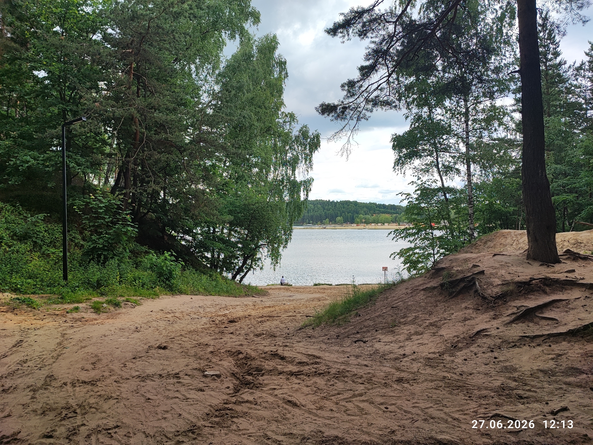 An eye-level, medium shot captures a sandy path leading down to a tranquil lake, framed by lush green trees and a cloudy sky. The path, marked with tire tracks, appears to be a natural trail. On the lake's shore, a lone figure sits facing the water, contemplating the expansive view. A red and white sign is visible in the distance. The foreground is dominated by a sandy incline with exposed roots, suggesting a natural, somewhat rugged landscape. The image, dated June 27, 2026, at 12:13 PM, likely depicts a public recreation area in Lytkarino, Russia, given its natural setting and potential for leisure activities.