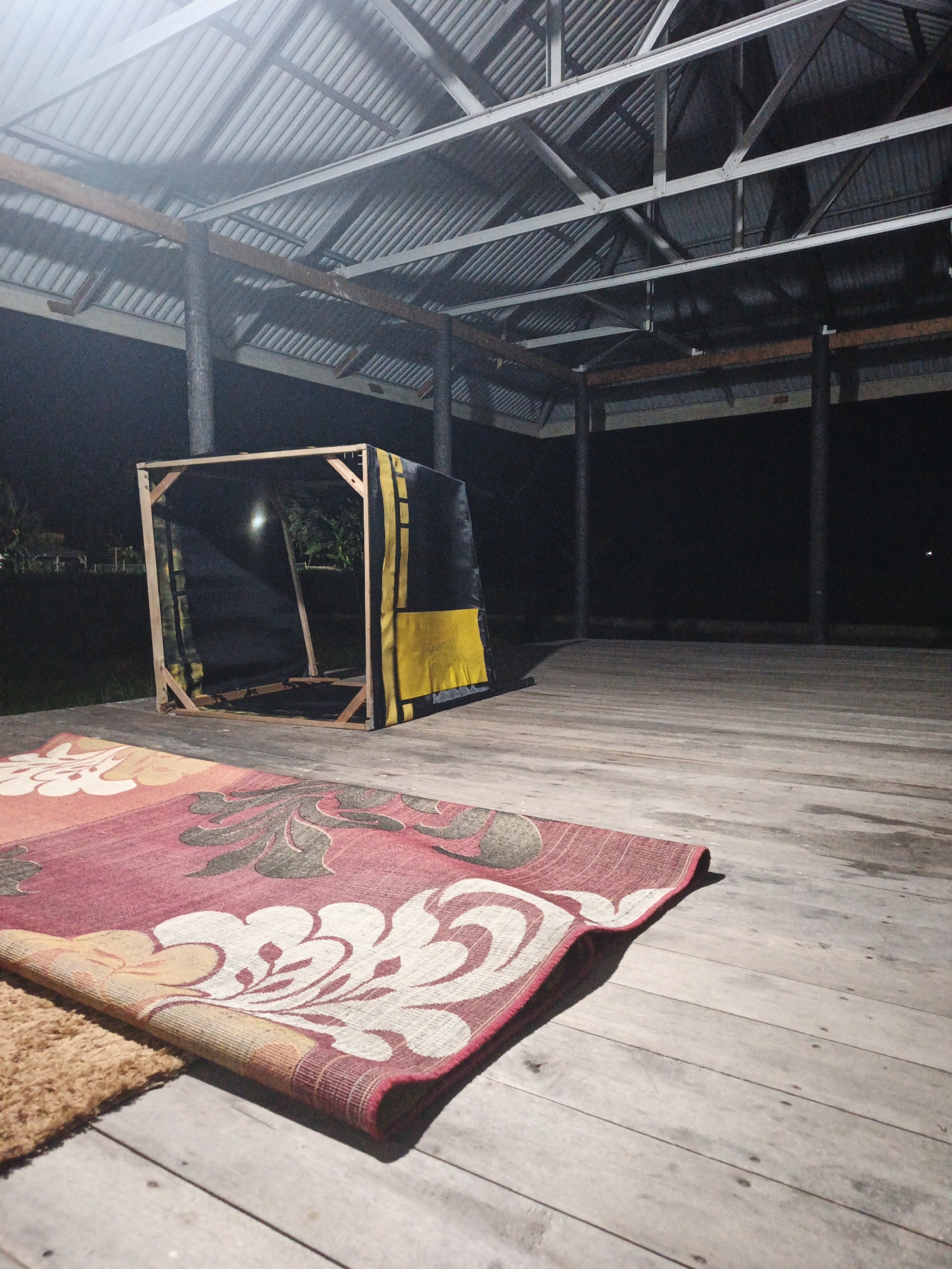 The image depicts an open-air structure with a corrugated metal roof supported by wooden posts and beams. A wooden-framed structure covered with black and yellow fabric is situated in the middle of the floor. Two rugs are laid out on the wooden floorboards. The scene is dark, suggesting it is nighttime, with some artificial lighting casting shadows. The location is identified as Kelapapati, Indonesia.