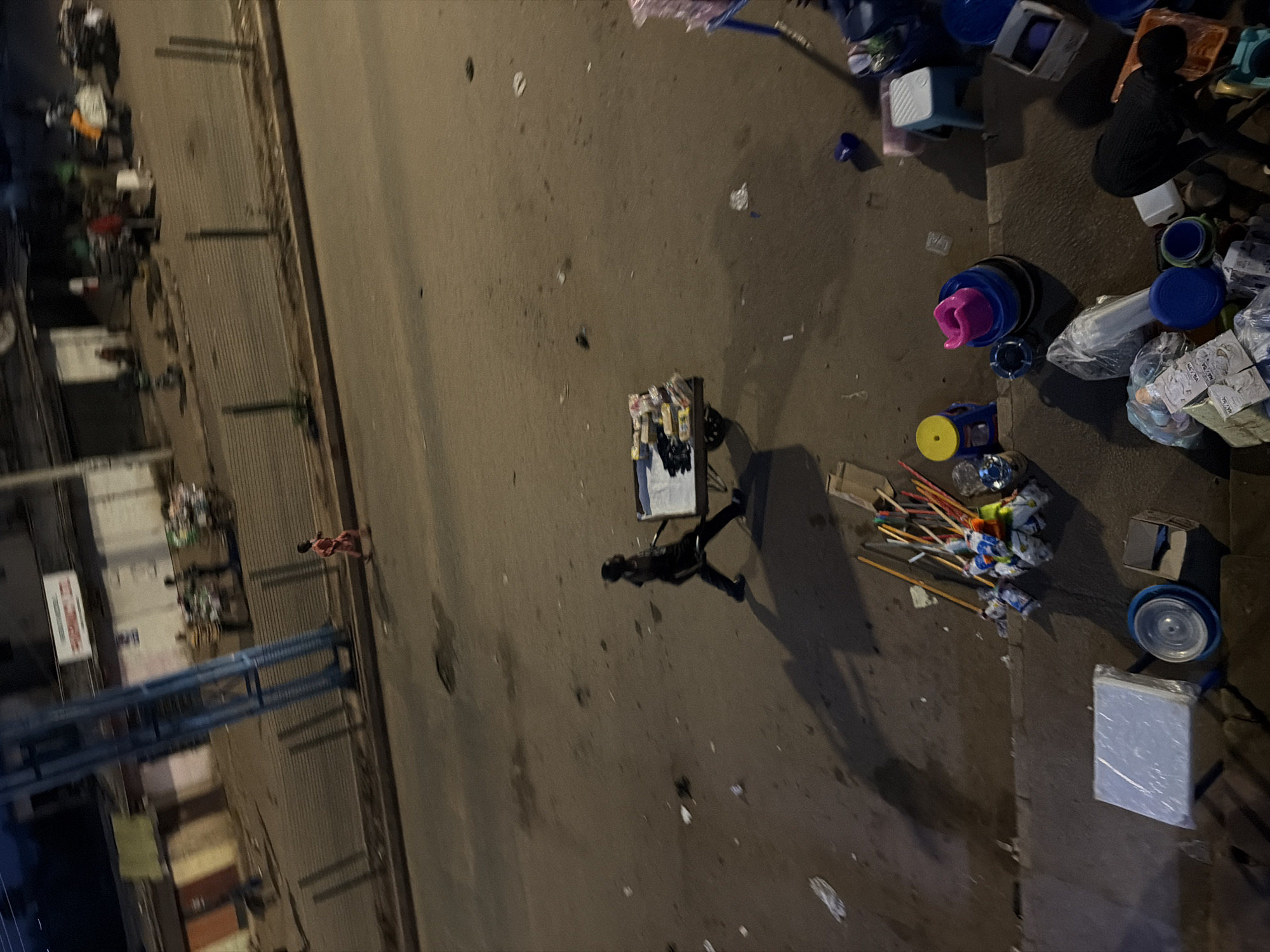 In Bukuru, Nigeria, a child in a red garment runs along the side of a dusty road at dusk. In the middle of the road, a person pushes a cart laden with goods. The scene is characterized by its grimy asphalt, scattered litter, and a few blurry figures in the background walking past storefronts. In the foreground, an assortment of cleaning supplies, including brooms and mops, are piled near what appears to be a makeshift stall, with stacks of plastic containers and boxes. The lighting suggests it is either early morning or late evening, casting long shadows across the scene.