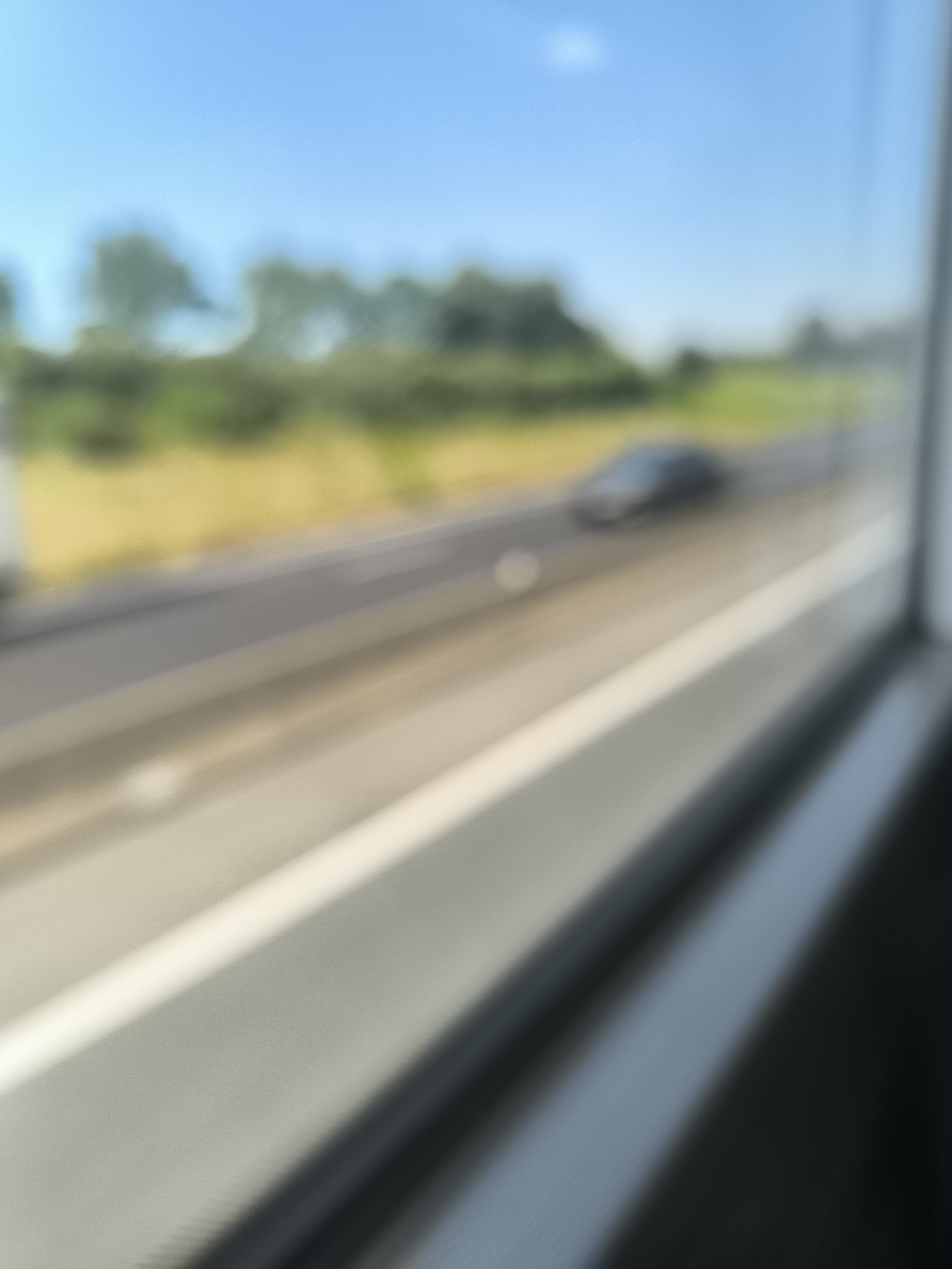 The image shows a blurry view from a window, likely of a moving vehicle. Outside, a highway runs parallel to a field or grassy area with trees in the background. A car is visible on the highway. The sky is blue. The location, Saint-Chamond, France, is provided context but is not explicitly visible in the image.