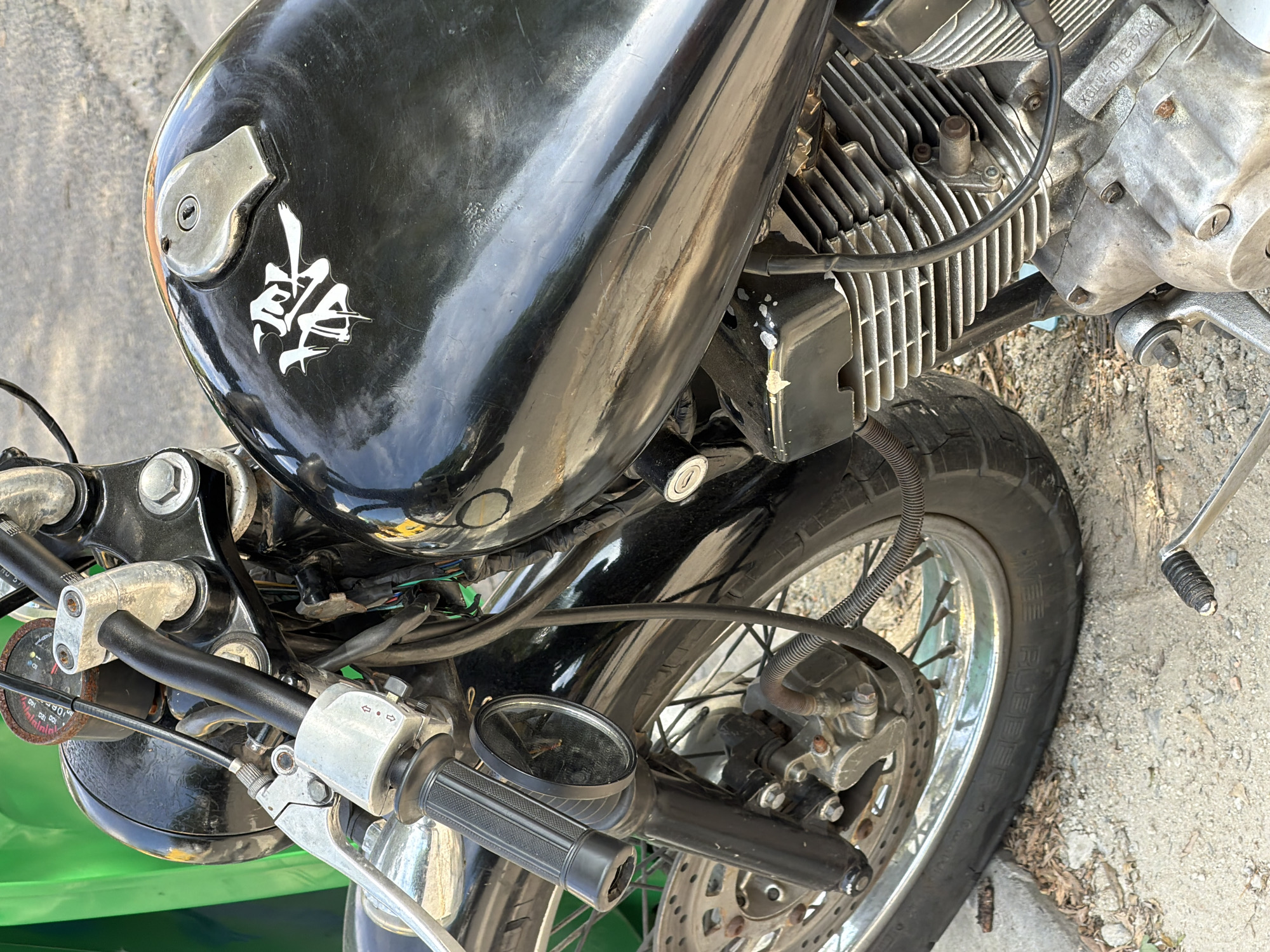A close-up, eye-level shot shows a partially visible motorcycle. The motorcycle features a black gas tank adorned with a white decal featuring an Asian script. The tank shows significant wear with scratches visible on its surface. To the right of the tank is a weathered engine with prominent cooling fins. The front wheel with a disc brake is also visible. The motorcycle's handlebars are equipped with standard controls and a side mirror. A green section of the motorcycle frame can be seen at the lower left. The bike rests on a concrete surface, potentially a sidewalk or road. There are no people visible. The location, Đà Nẵng, Vietnam, cannot be confirmed from the image.