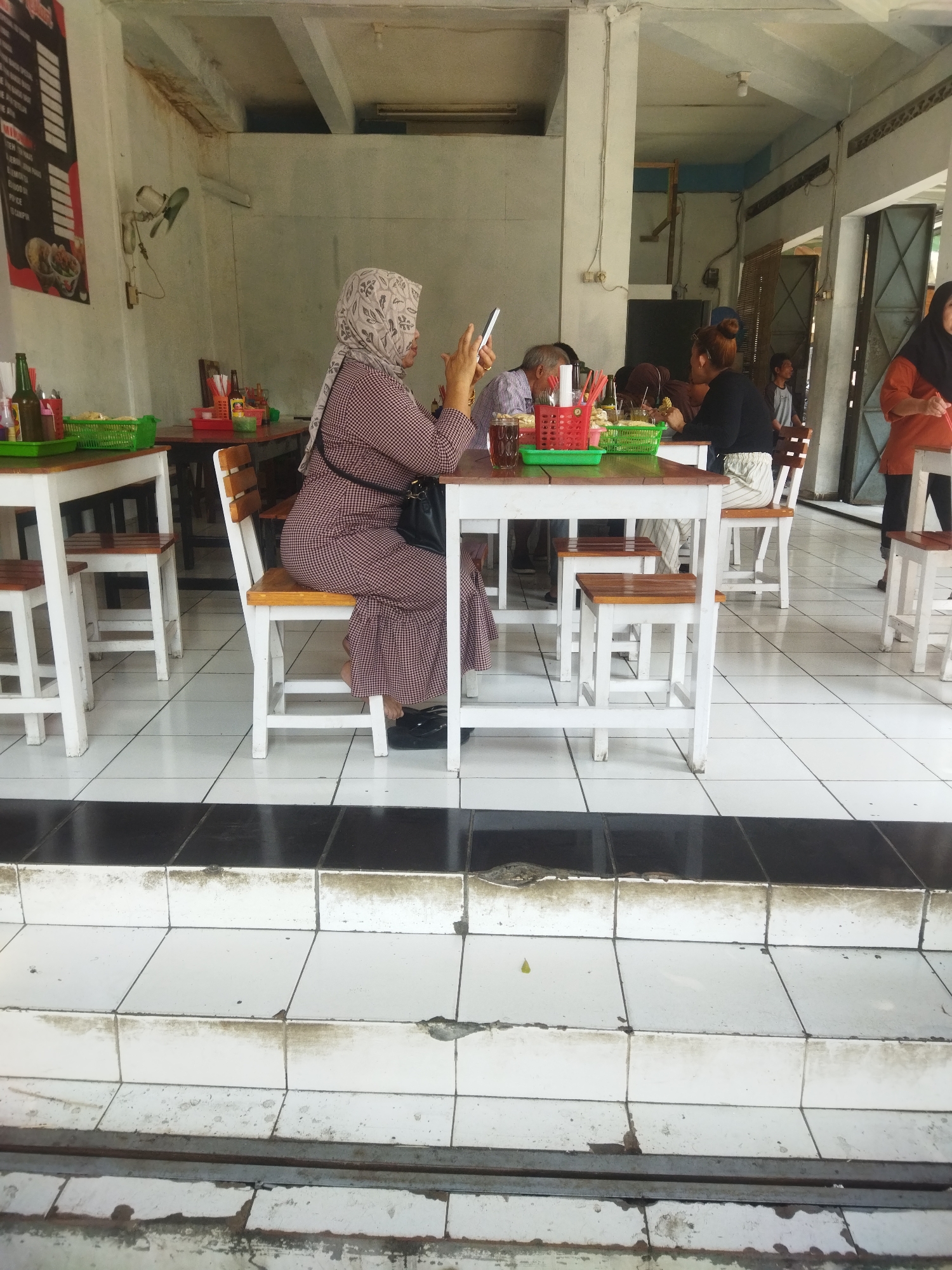 A medium shot captures an outdoor seating area of a restaurant in Wonogiri, Indonesia, with a focus on a woman sitting at a table and checking her phone. The woman, wearing a patterned hijab and a plaid dress, is seated on a white chair with a wooden seat. She is positioned at a white table with a dark wooden top, where other diners are also present, though less clearly visible. In front of her on the table are a phone, a small black handbag, and various dining items. The environment appears to be a covered outdoor space, with white tiled flooring. The background shows other tables and chairs, as well as architectural elements typical of a casual Indonesian eatery, including exposed beams and wall partitions. The overall scene conveys a relaxed atmosphere of people dining and interacting.