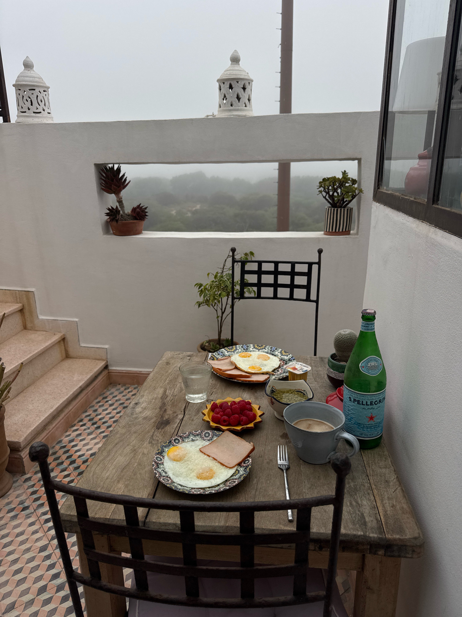 The image depicts a breakfast setting on a rustic wooden table placed on a terrace, likely in Essaouira, Morocco, given the architectural style and surrounding environment. The atmosphere is overcast, with a light fog obscuring the distant landscape, which appears to be a mix of vegetation and possibly distant buildings.
On the table, two plates of breakfast are arranged. One plate contains two fried eggs and a slice of ham, while another plate holds what appears to be a slice of ham and two fried eggs. A small dish of raspberries is also present, along with a small container of what could be yogurt or jam. A glass of water, a cup of coffee, and a bottle of S.Pellegrino sparkling water are also on the table, alongside a fork.
The terrace itself is enclosed by white walls, with decorative white lanterns mounted on the parapet. A set of stone stairs with a patterned tile runner leads up to the terrace on the left side of the image. In the background, a small potted tree and a potted succulent are visible, adding a touch of greenery to the scene. A black metal chair is pulled up to the table, and another chair is visible in the background. The flooring on the terrace is a patterned tile in shades of pink, orange, and gray. A small cactus plant is situated next to the S.Pellegrino bottle. A window with a dark frame is visible on the right side of the image, through which a red ceramic object can be seen. The overall impression is one of a tranquil, slightly misty morning breakfast on a Moroccan rooftop terrace.