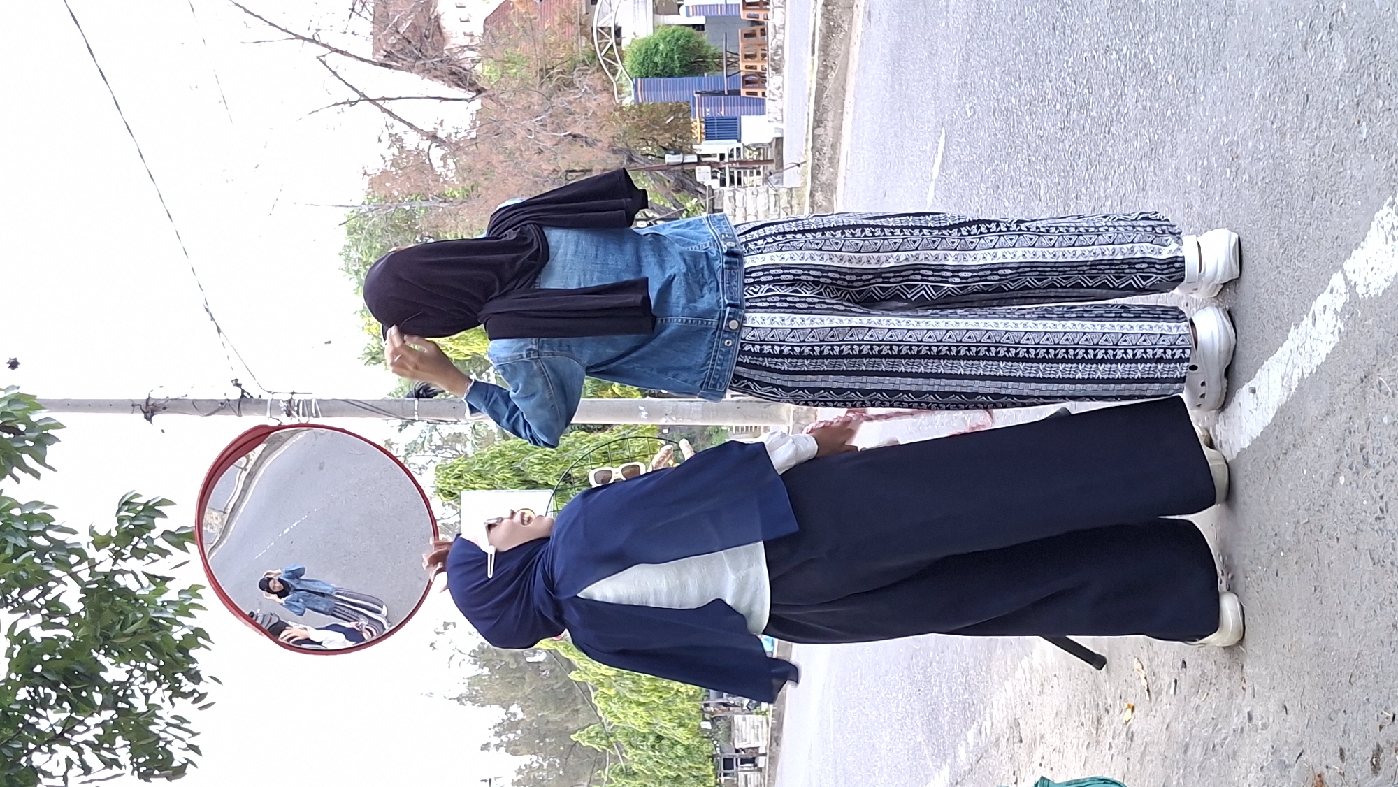 In Lhokseumawe, Indonesia, two young women stand near a convex mirror mounted on a pole. The woman on the left wears a dark blue hijab, a white blouse, and dark blue wide-leg pants, along with white slip-on shoes. The woman on the right sports a black hijab, a denim jacket, and patterned pants in black, white, and blue, also wearing white slip-on shoes. Both are facing away from the camera, with the woman on the right adjusting her hijab. The mirror reflects the street and the woman on the left, appearing smaller. The environment is an urban street with a paved road, a sidewalk, trees, and buildings in the background.