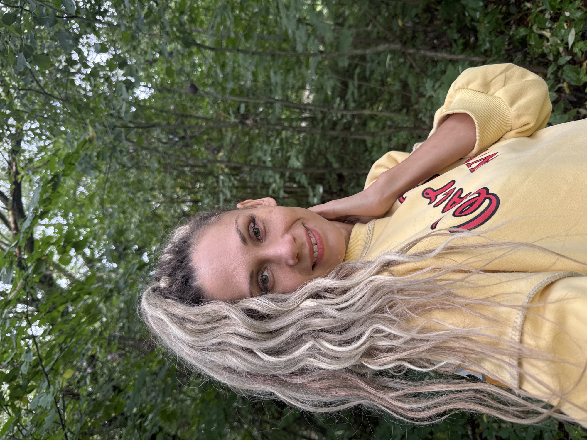 In the city of Rusanovo, Russia, a woman with long, blonde, wavy hair stands in a wooded area. She is wearing a yellow sweatshirt with red lettering. The woman is looking at the camera and smiling, with her hand resting on her neck. The background is filled with lush green trees.
