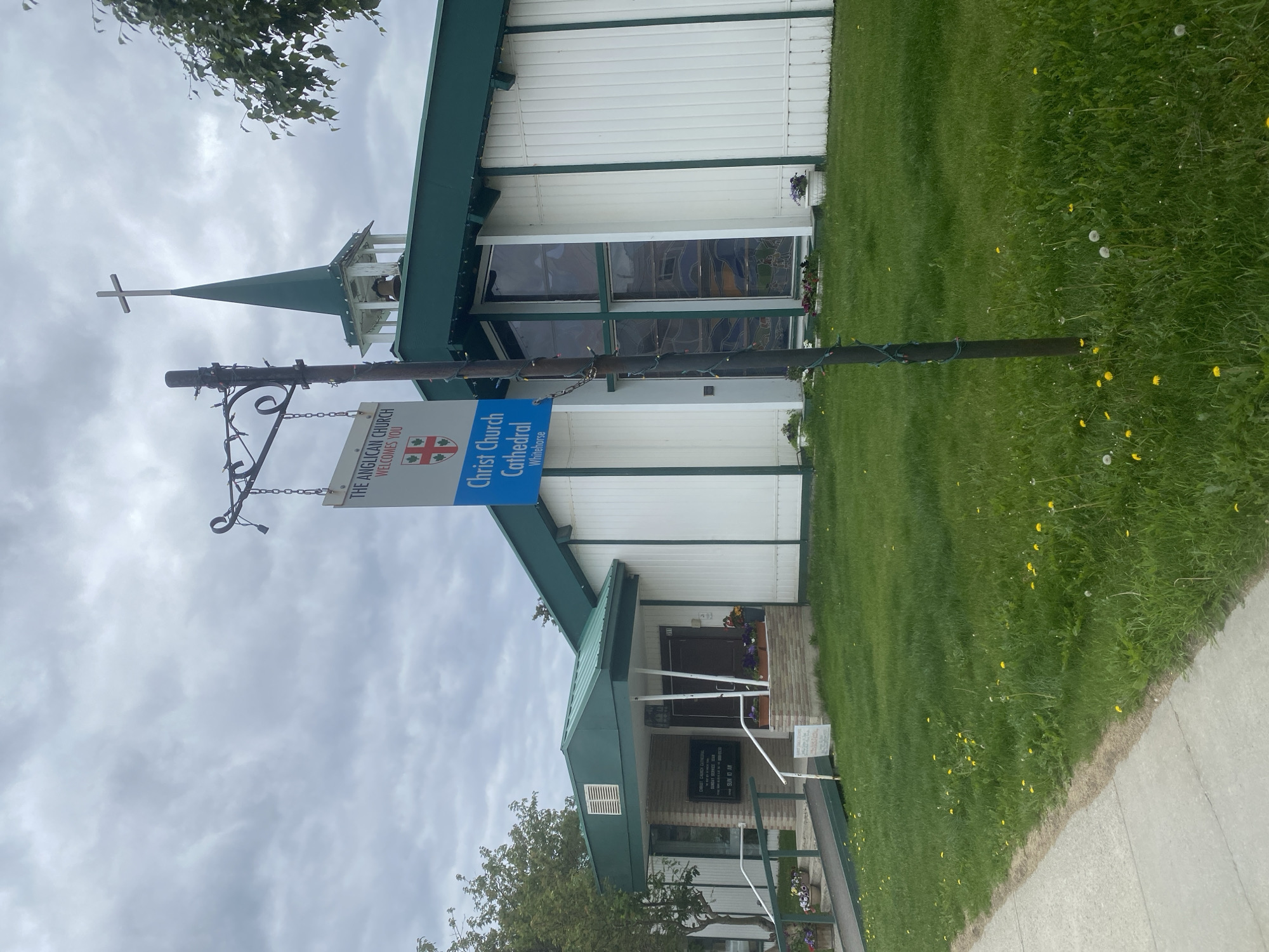 The image shows the exterior of Christ Church Cathedral, located in Whitehorse, Canada. The building is a modest structure with white vertical siding and a green roof. A steeple with a cross rises from the roof, and a bell is visible within the steeple. A sign in front of the building reads "THE ANGLICAN CHURCH WELCOMES YOU" and "Christ Church Cathedral Whitehorse." The church has large stained-glass windows on the side. The grounds are covered in grass with scattered dandelions and some potted flowers near the entrance. A sidewalk runs along the left side of the image. The sky is overcast. There are no people visible in the image.