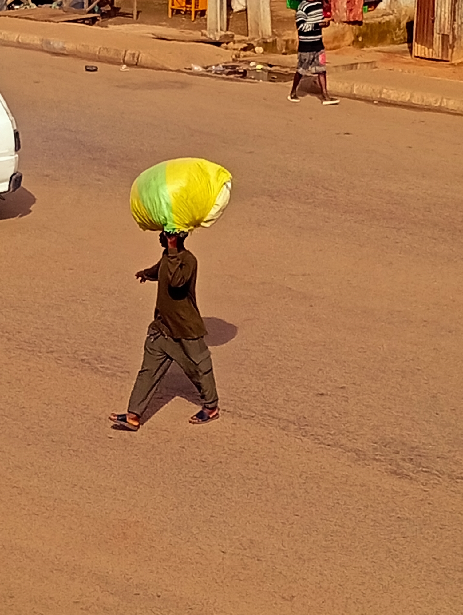 A person walks across a dusty, paved road in Bukuru, Nigeria, carrying a large, green and yellow bundle on their head. The person is wearing a dark long-sleeved shirt, dark pants, and sandals. The road is lined with a curb and sidewalk, with buildings and some debris visible in the background. Another person in the distance, wearing a striped shirt and patterned shorts, is also walking on the sidewalk. A white vehicle is partially visible on the left side of the image. The overall scene depicts a typical day in a Nigerian city.