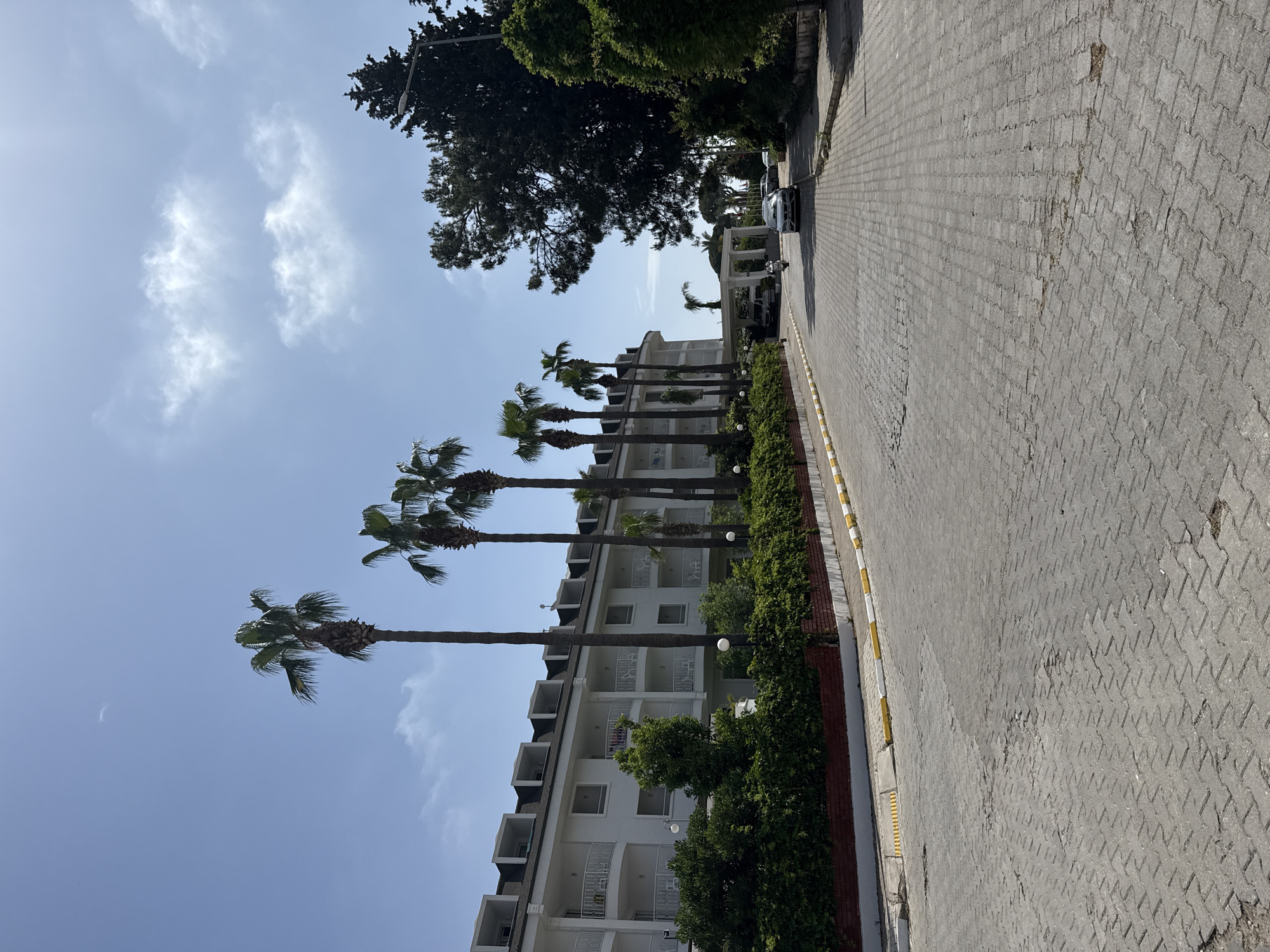 The image shows a street scene in Kemer, Turkey. A multi-story, white building with balconies and palm trees lining its front stands alongside a paved road. The sky is blue with scattered clouds. There are various vehicles parked further down the street. Green foliage borders the building and lines the road.