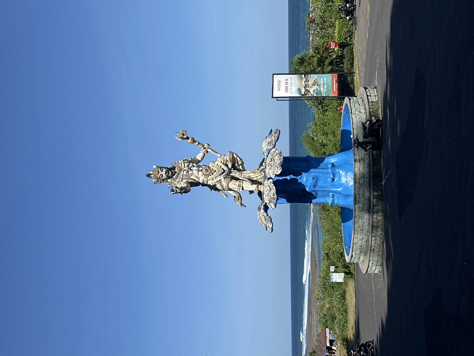 The image depicts a stone statue of a deity figure, possibly Hindu or Balinese, located in Kuta Selatan, Indonesia. The statue is positioned atop a blue and grey pedestal near a paved road and a coastline. A sign for "Roosterfish" restaurant is visible in the background. The sky is clear and blue. A glimpse of the ocean is visible. A sign with "OUT" is seen. There are motorbikes parked nearby.
