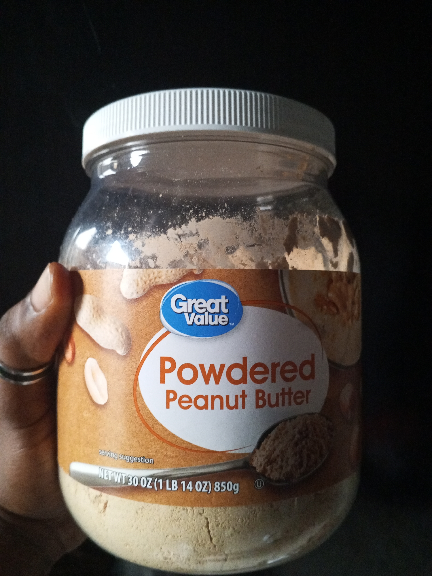A hand wearing two silver rings is holding a clear plastic jar of Great Value Powdered Peanut Butter. The jar is about two-thirds full of a light brown powder. The label on the jar is predominantly orange and white with blue lettering. The words "Great Value" are in a blue oval, and below that, "Powdered Peanut Butter" is written in large orange letters. A silver spoon filled with the powdered peanut butter is resting on the label. In the background, a dark, out-of-focus area suggests an indoor setting. The image is a close-up shot focused on the jar of peanut butter. There is no information in the image to indicate a location in Ashata, Nigeria.