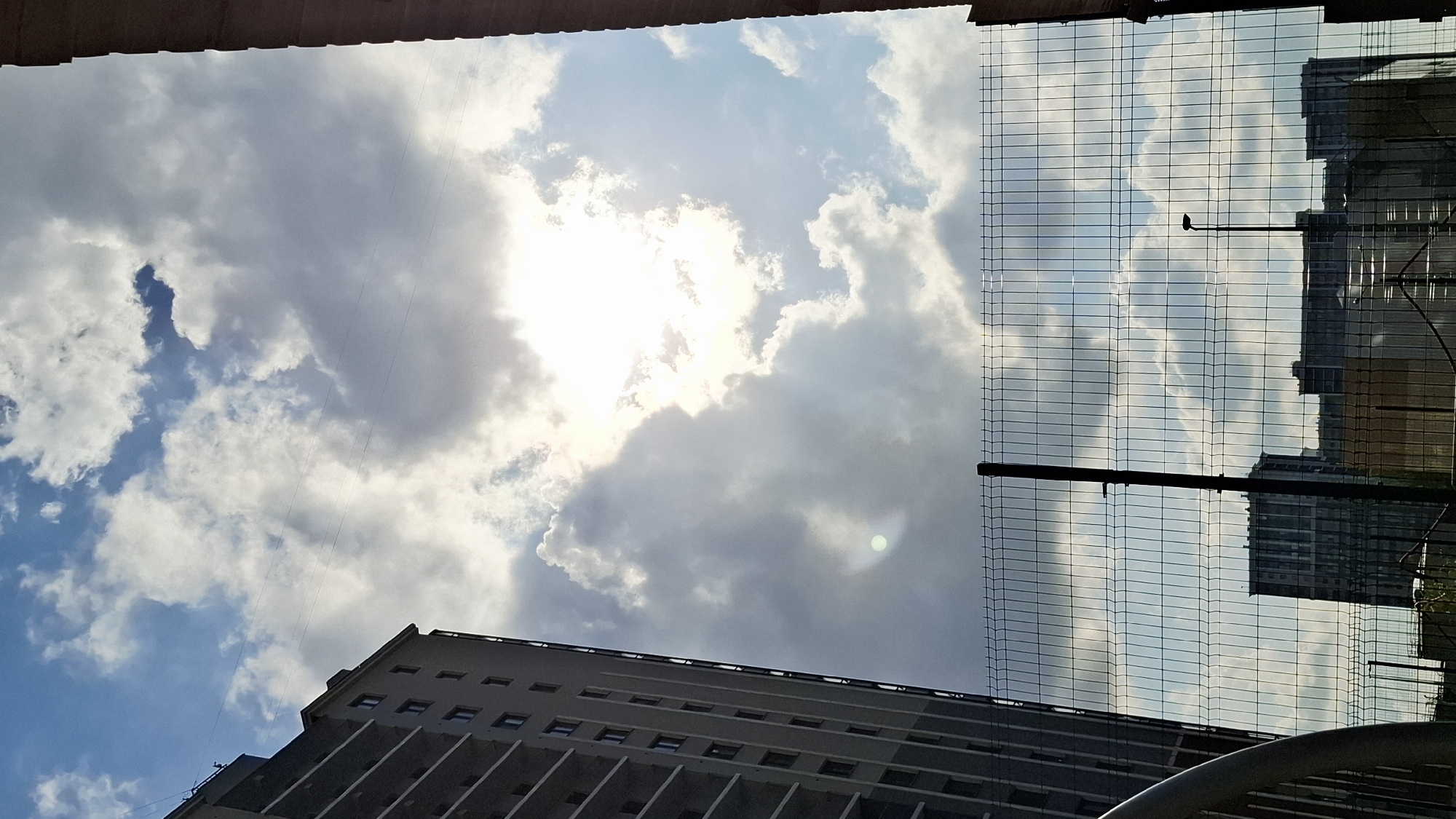 A low-angle shot looking up at a cloudy sky with the sun obscured by clouds. At the bottom of the frame, a large grey building with multiple rows of windows is visible. To the right, a wire mesh fence is present, partially obscuring other buildings in the background. A dark structure, possibly a pole, extends upwards through the fence. The photograph might have been taken in Saint Petersburg, Russia.