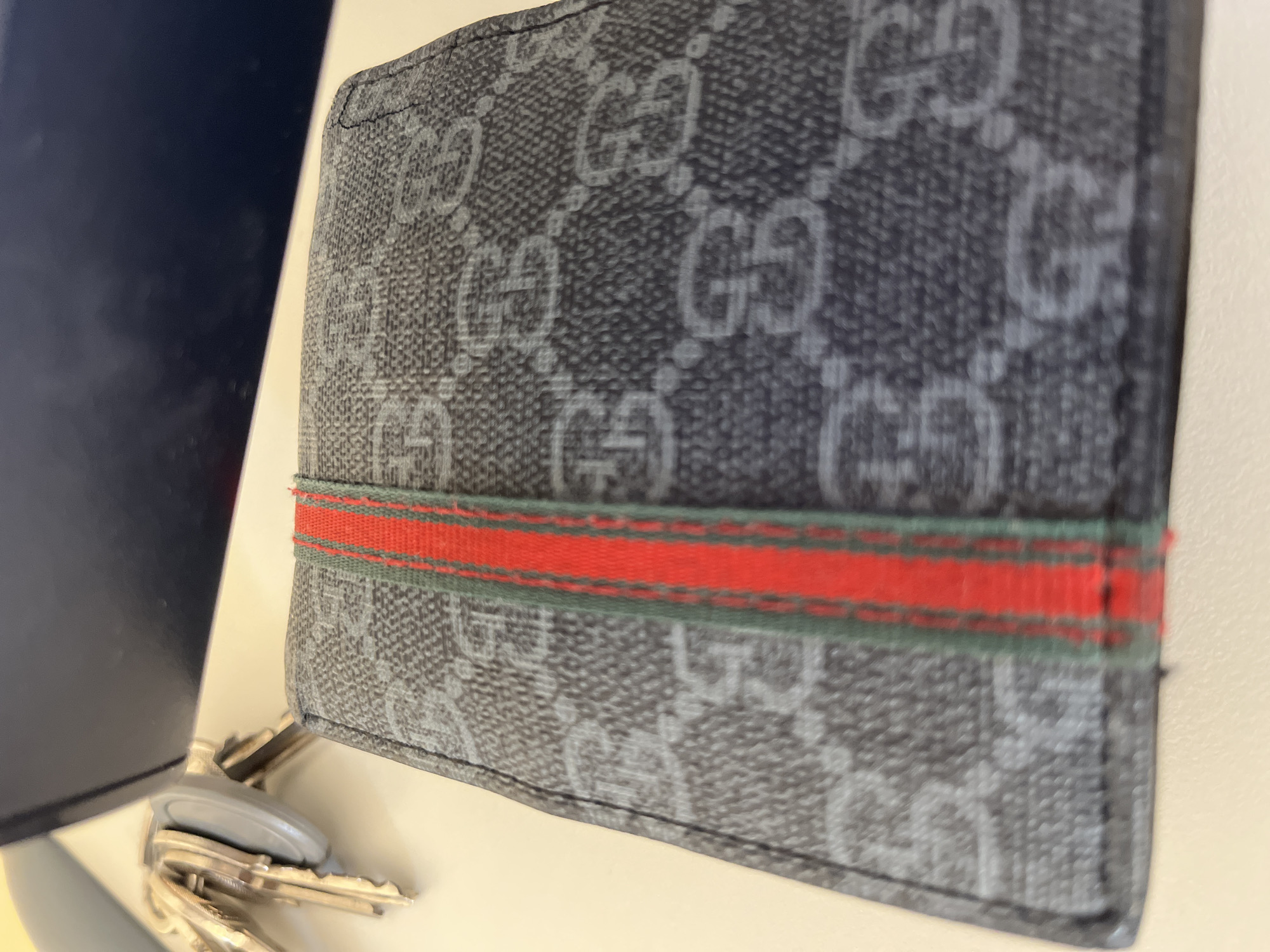 The image presents a close-up, slightly angled view of a wallet and a set of keys on a light-colored surface. The central subject is a bi-fold wallet, predominantly dark gray or black, adorned with a repeating pattern of lighter gray interlocking 'G' logos, characteristic of the Gucci brand. A prominent red and green striped web band runs horizontally across the middle of the wallet. The material of the wallet appears textured, possibly a coated canvas.
In the lower-left corner, a small cluster of metallic keys is visible, attached to a gray keyfob. Partially obscuring the keys on the far left is a dark blue or black object with a smooth, somewhat reflective surface. The setting appears to be an indoor environment, likely a desk or table, given the static arrangement of personal items. No individuals are present, and no particular activity is depicted. The lighting is soft and even, highlighting the details of the wallet's pattern and the vibrant colors of its striped accent. The visible text is the recurring 'GG' logo across the wallet's surface.
