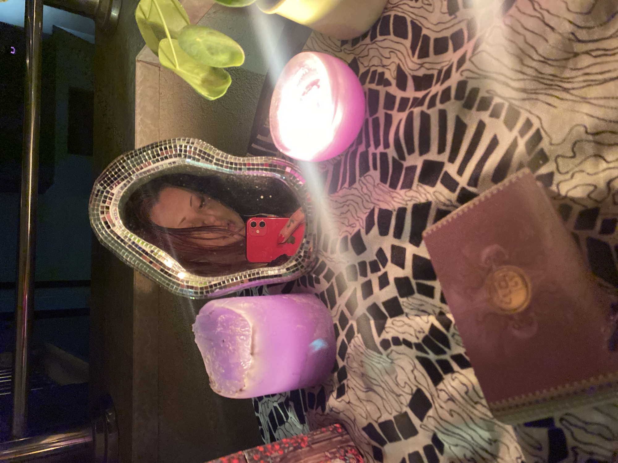 This image depicts a tabletop arrangement in Boreti, Montenegro, illuminated primarily by candlelight. Centrally positioned is an irregular-shaped mirror framed with small square mosaic tiles, reflecting a female subject. The subject's face is partially obscured by dark hair and tilted downwards, while her right hand, holding a red smartphone with multiple camera lenses, is clearly visible in the reflection, indicating she is taking the photograph. Her fingernails are painted red.
On the left, a large, irregular-shaped purple candle, showing signs of melting, stands next to a small, decorative, reddish object. To the right of the mirror, a spherical pink candle emits a bright light, suggesting it is actively burning, with lens flares visible emanating from it. Further to the right, a third, lighter-colored cylindrical candle in a beige holder is partially visible. The objects rest on a fabric surface featuring a black and white geometric pattern. In the foreground, a maroon-colored book with a gold circular emblem on its cover is placed. Green leaves from a plant are visible in the upper right background. The background is dark and out of focus, showing what appears to be a metallic railing or bar in the upper left, suggesting an indoor or covered outdoor setting with dim ambient lighting.