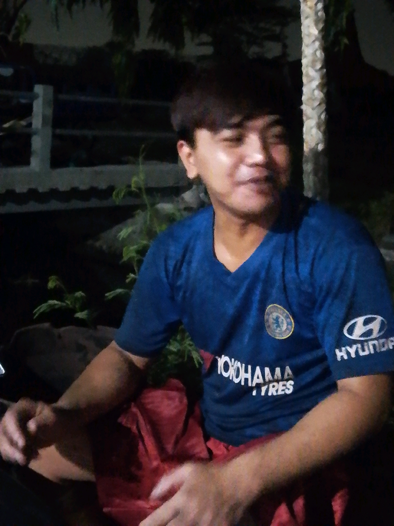 A man is sitting outdoors at night, illuminated by artificial light. He is wearing a blue t-shirt with logos on it, including a circular emblem on the left chest and a Hyundai logo on the right sleeve. He has short, dark hair and is looking slightly off to the side, with a faint smile. His hands are resting on his lap, which is covered by a red cloth. The background is dark and shows some greenery, including what appears to be a palm tree to the right of the man. Behind him, there are structures that suggest a fence or railing. The overall impression is a casual, informal setting.