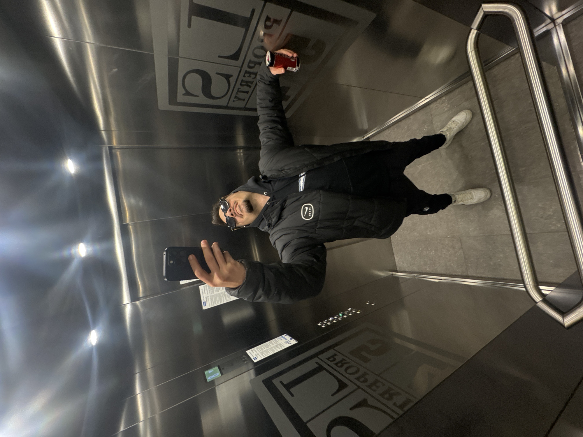A man is taking a selfie in an elevator. He is wearing a black puffer jacket, sunglasses, and white sneakers. He is holding a can in one hand and a smartphone in the other. The elevator has a brushed metal interior with a handrail. The buttons and signs inside the elevator are in Cyrillic, with the word "LOBERIA" visible, suggesting this is an elevator in Loberia, Sofia, Bulgaria.
