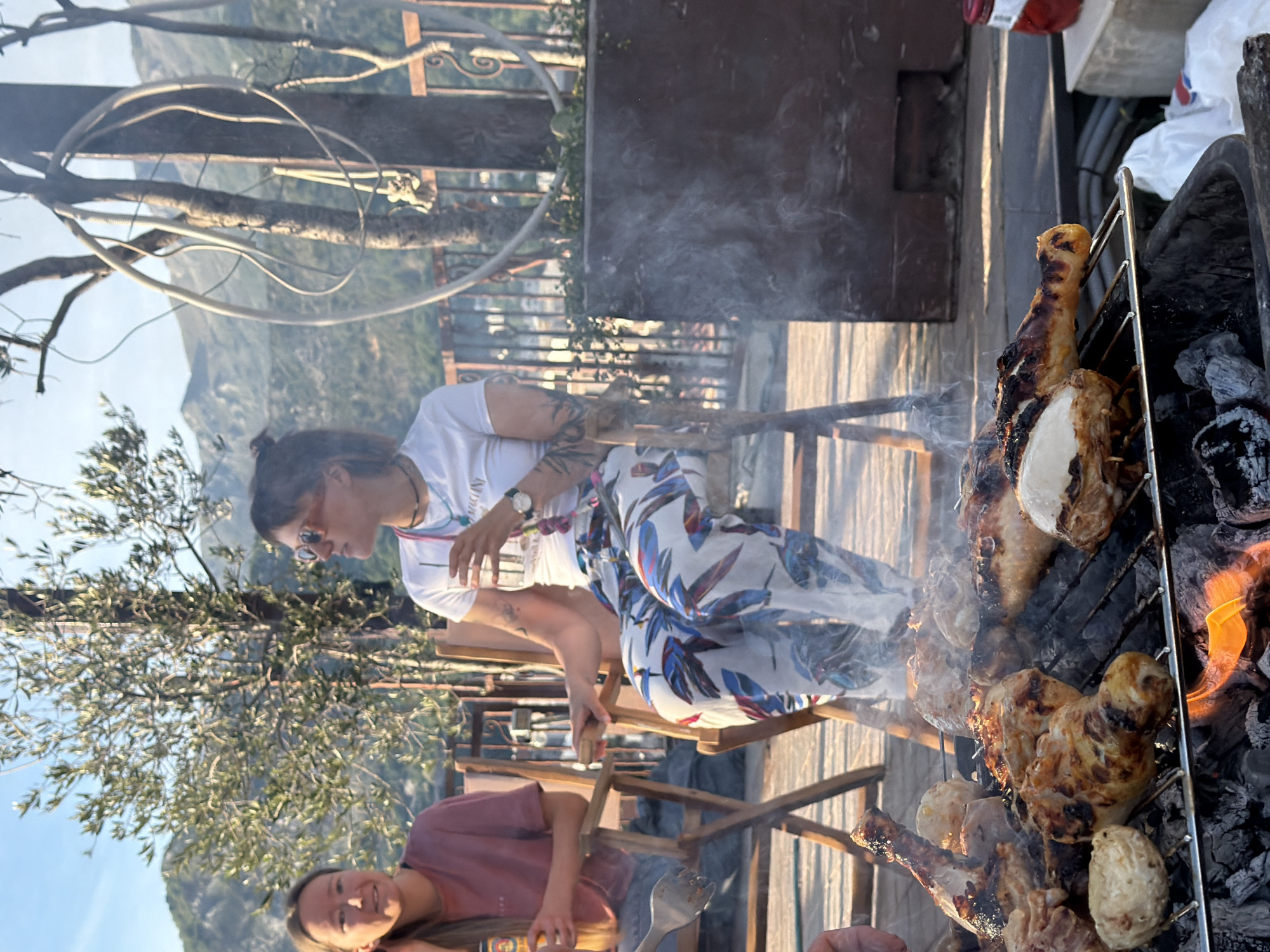 The image captures a scene of an outdoor barbecue, likely taking place on a patio or terrace, possibly in a residential area. In the foreground, there is a grill with pieces of chicken cooking over hot coals, producing smoke. A woman is grilling. Next to her, another woman is sitting in a wooden chair. The woman is wearing a white t-shirt and patterned pants. In the background, there is a scenic view of mountains and cityscape, suggesting the location might be in a hilly or elevated area.
The location is Budva, Montenegro.
